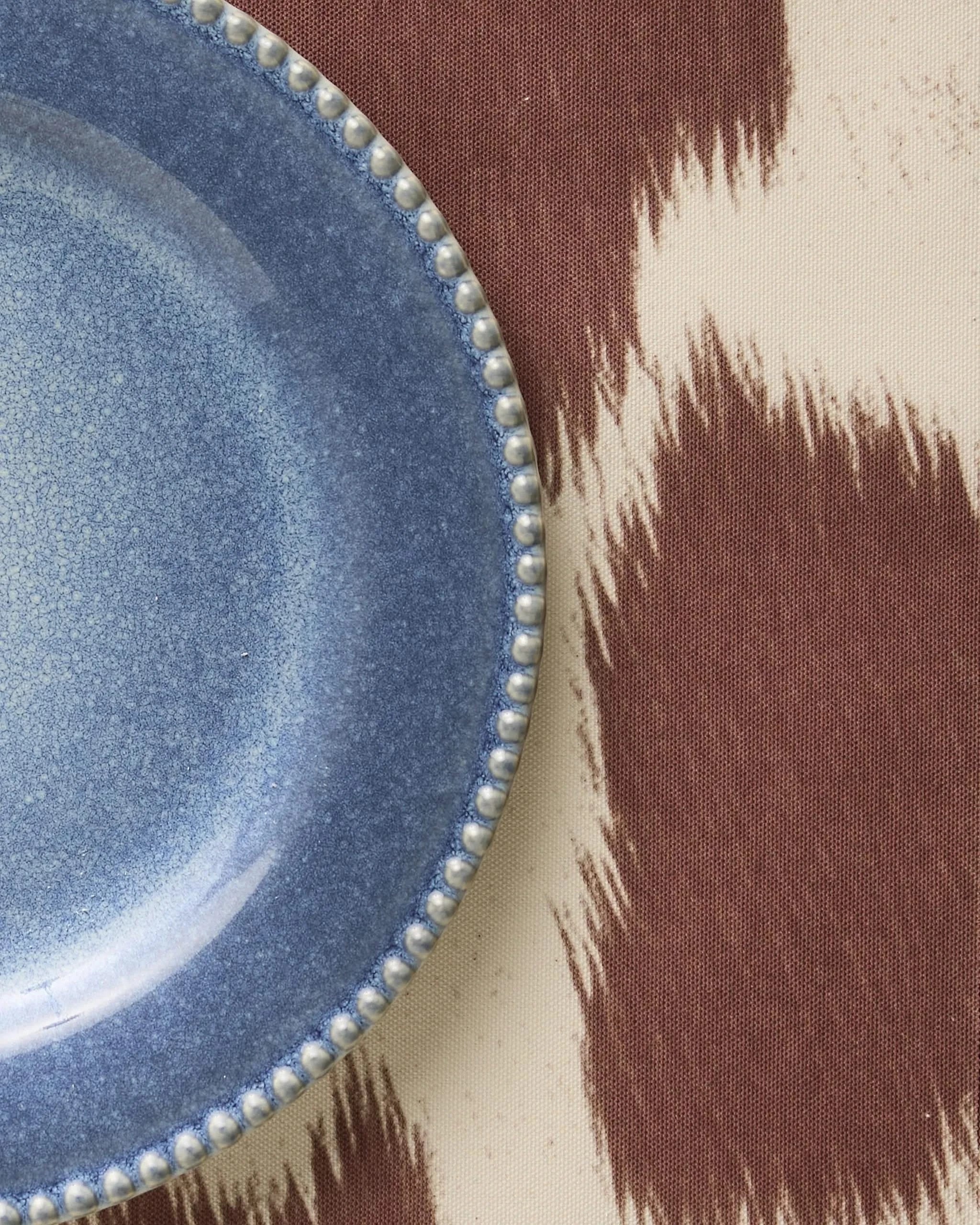 A close-up of a blue ceramic plate with a beaded edge, placed on brown and cream patterned fabric featuring Geometry’s Rodeo placemat.