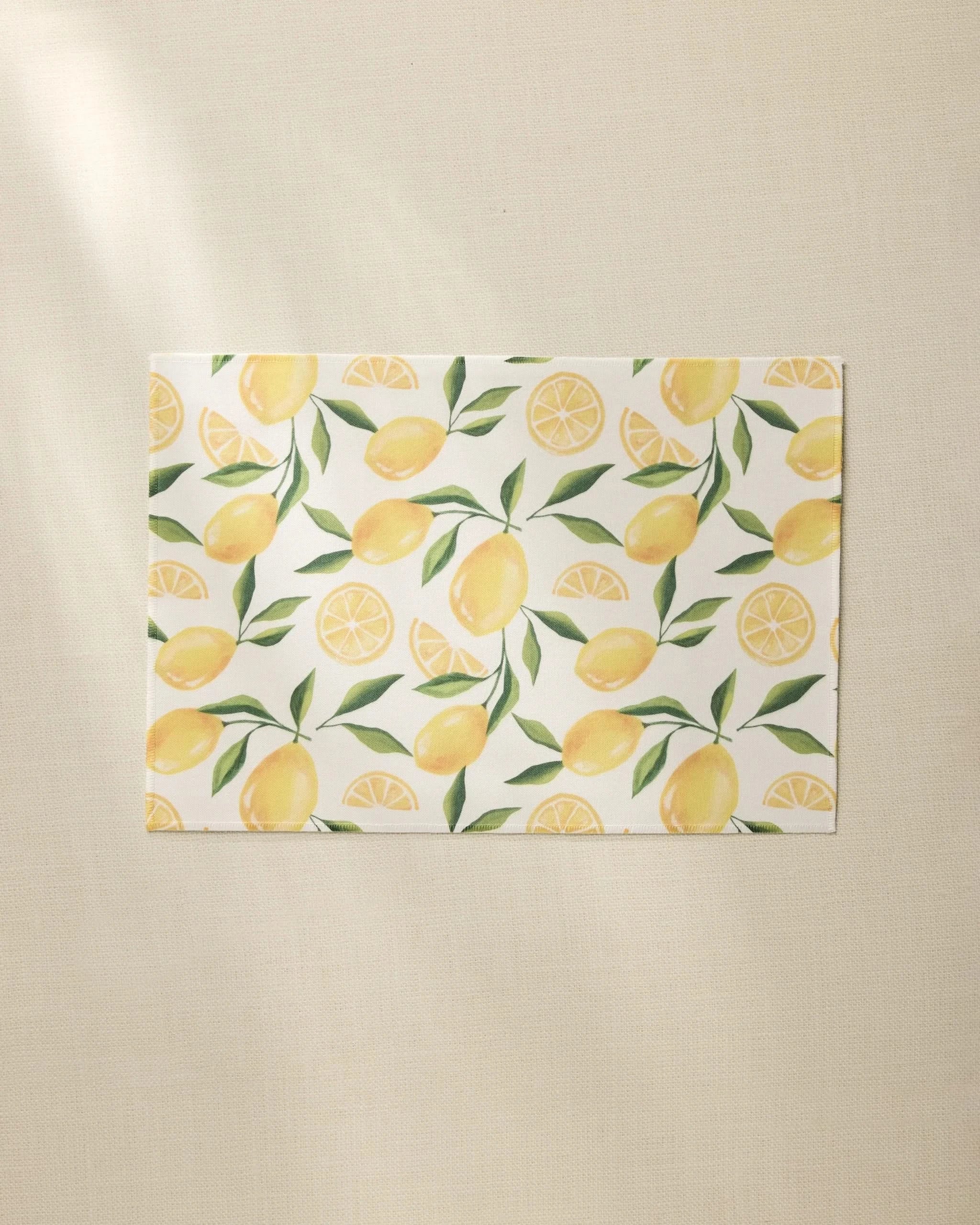 The Geometry Limoncello rectangular white recycled placemat features a pattern of whole lemons, lemon slices, and green leaves on a light beige textured surface.