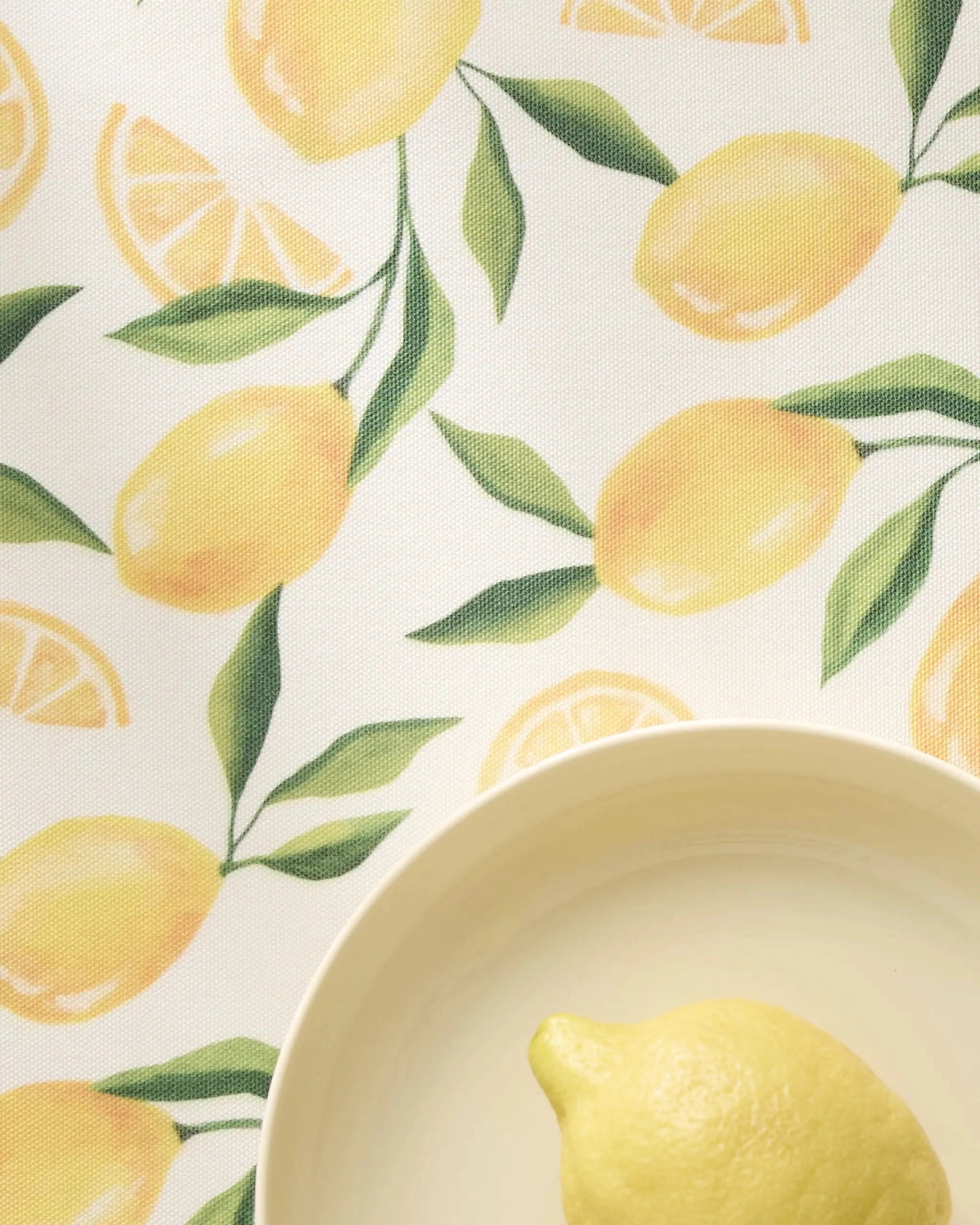 A whole lemon in a cream-colored bowl sits atop Limoncello table linens by Geometry, featuring lemon and leaf designs and durable placemats made from recycled materials.