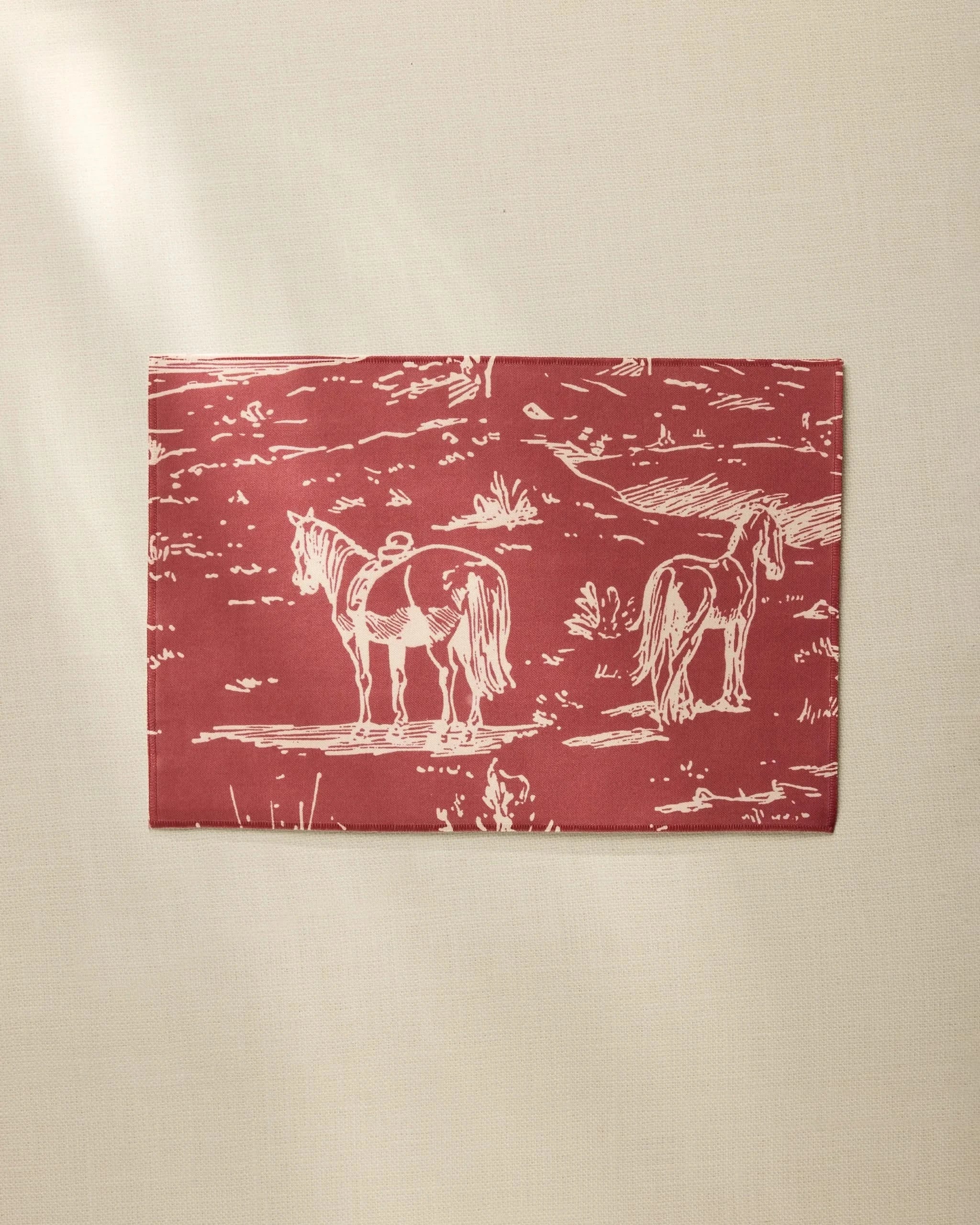 The Geometry Western Scene placemat is rectangular, red, and made from recycled materials, featuring a white line drawing of two horses in a field, shown on a beige surface.