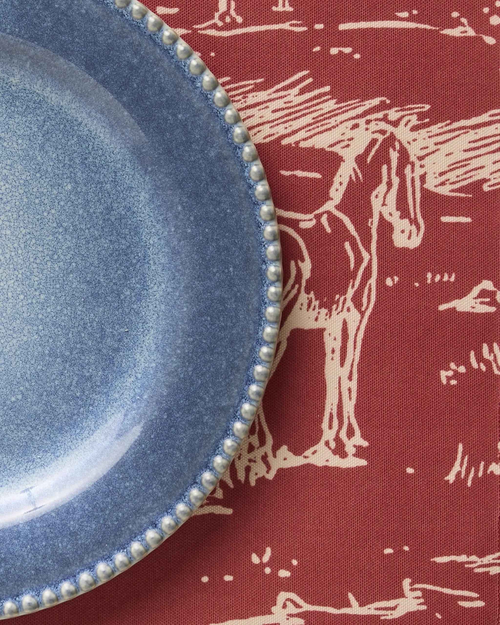 A blue ceramic plate with a beaded edge rests on the Geometry Western Scene placemat—a red, machine-washable tablecloth adorned with a white line drawing of a horse and landscape.