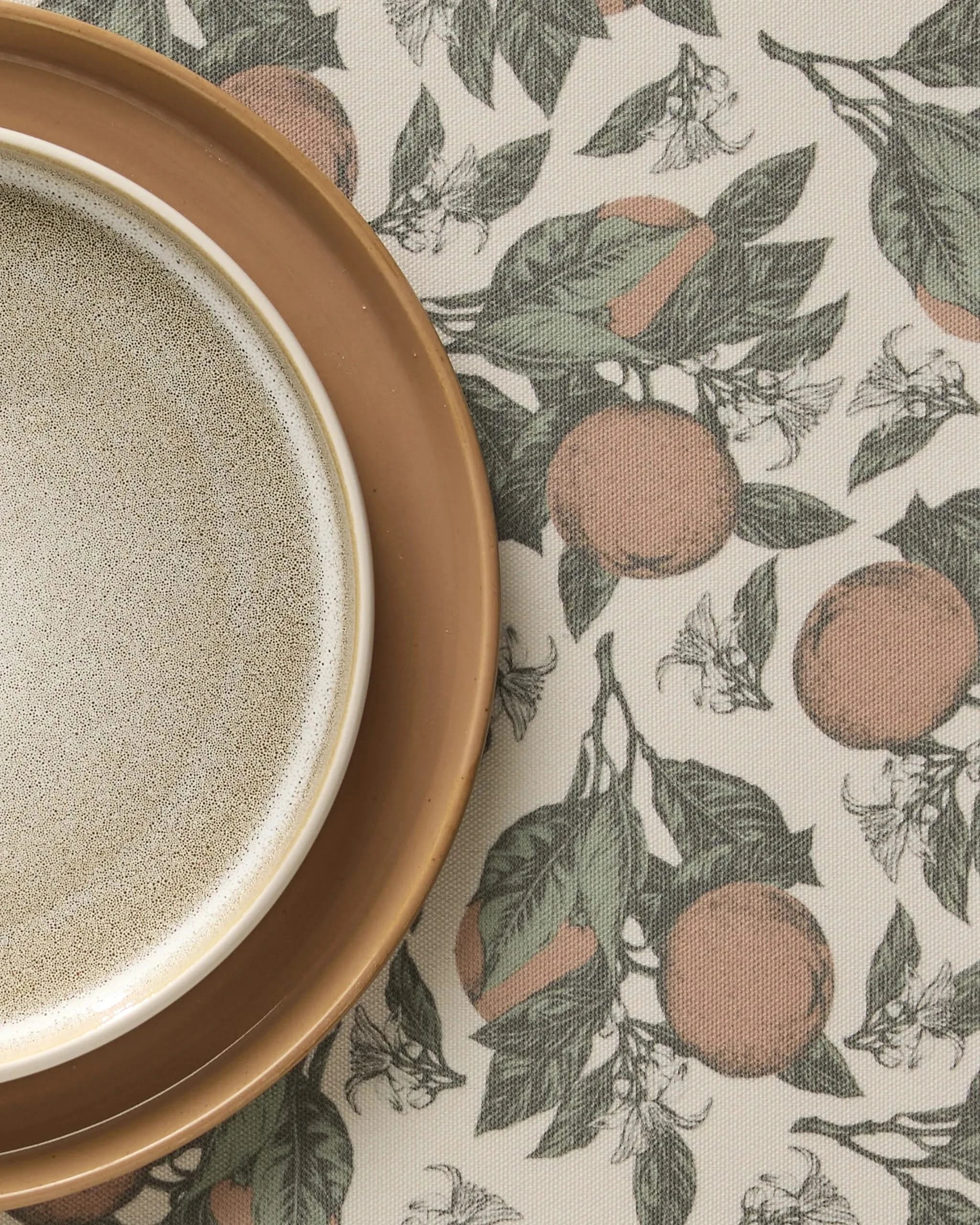 A close-up of the beige bowl on a brown plate is shown atop Geometry’s Valencia placemats, featuring a charming pattern of oranges, green leaves, and white blossoms. The placemats are machine washable.