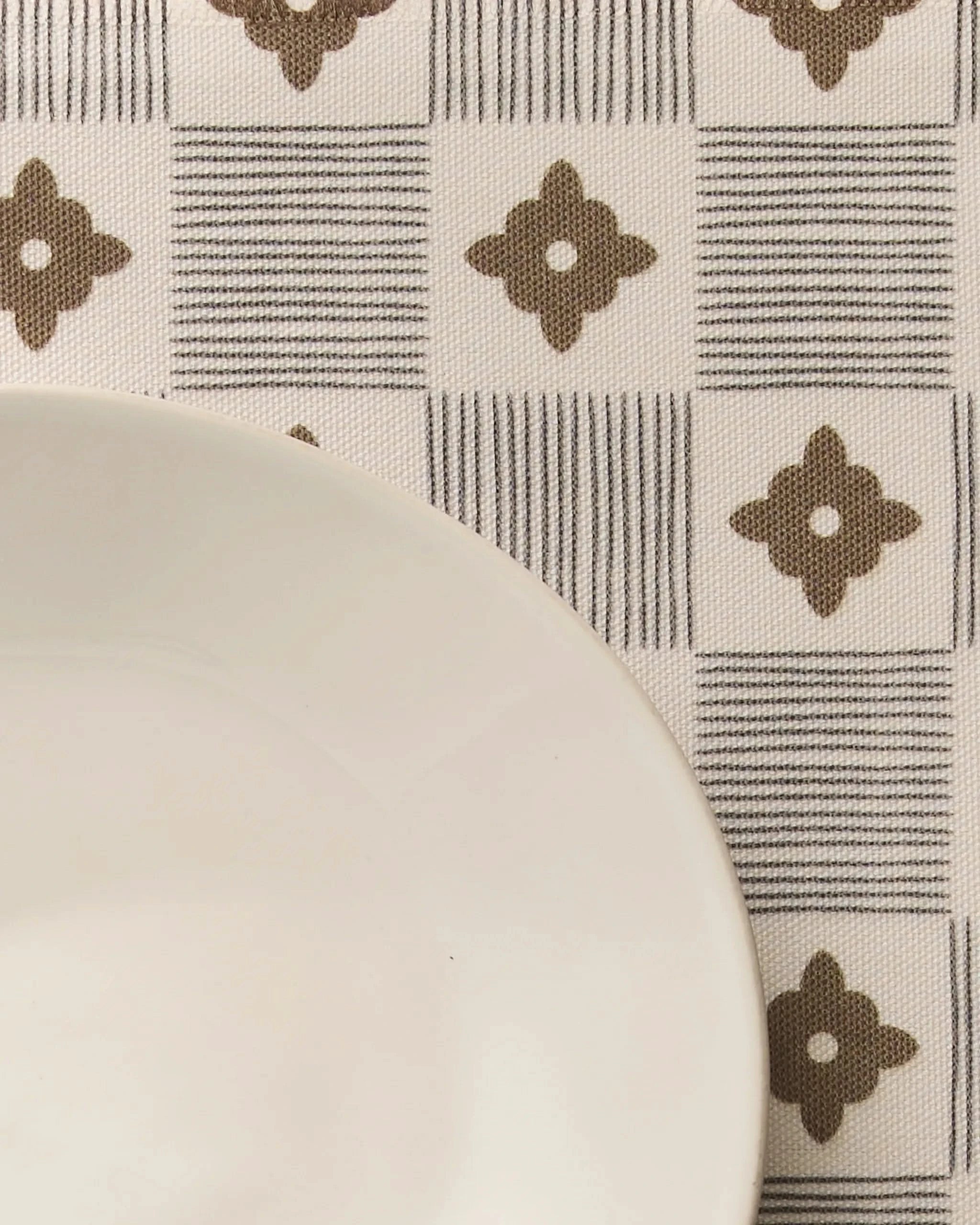 A close-up of Geometrys Legacy Block—an eco-friendly tablecloth with alternating striped squares and brown four-petal floral shapes, partly covered by a white plate in the lower left corner.