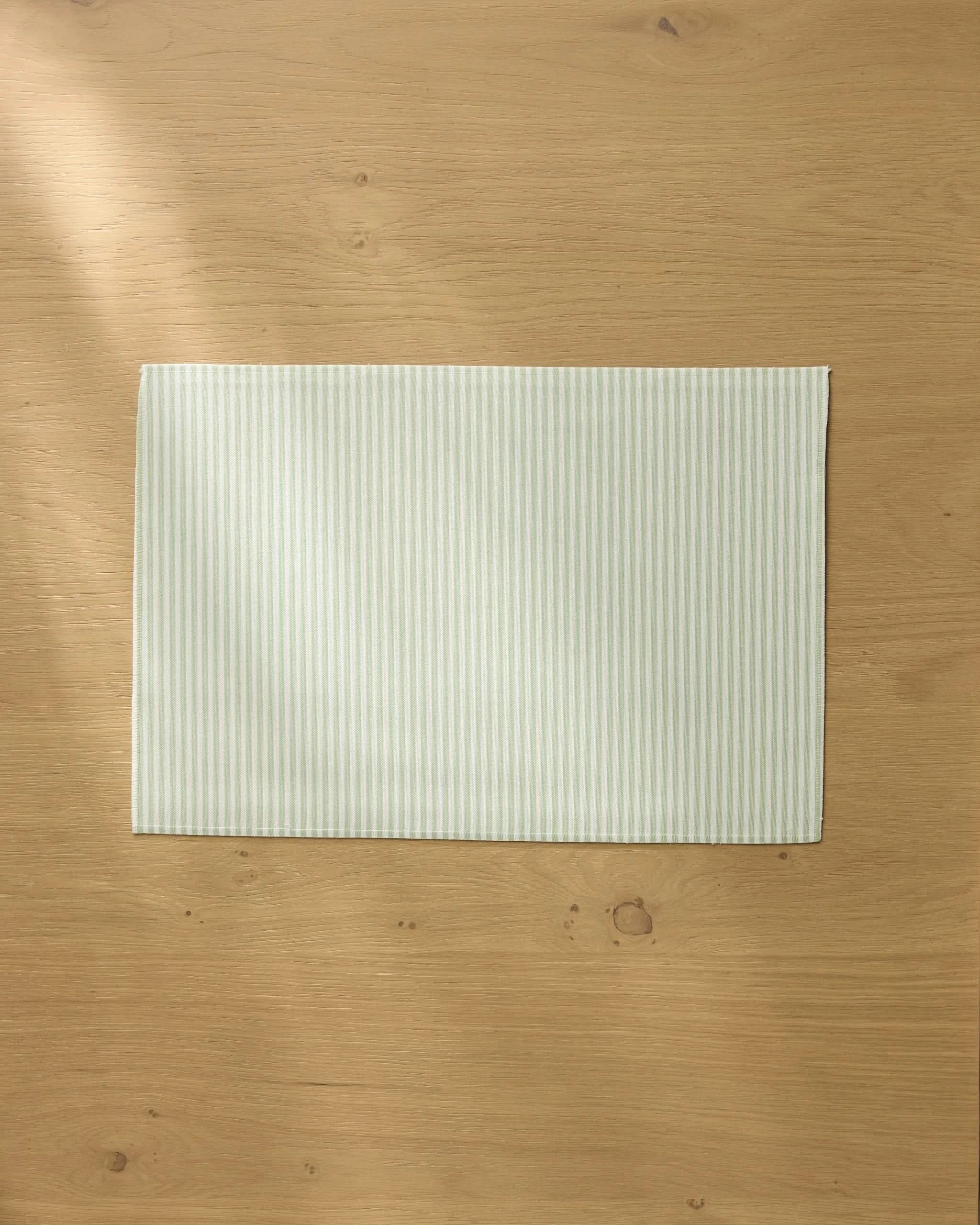 A pale green and white striped Seagrass placemat by Geometry rests on a light wooden table, with soft natural light casting gentle shadows across the surface.