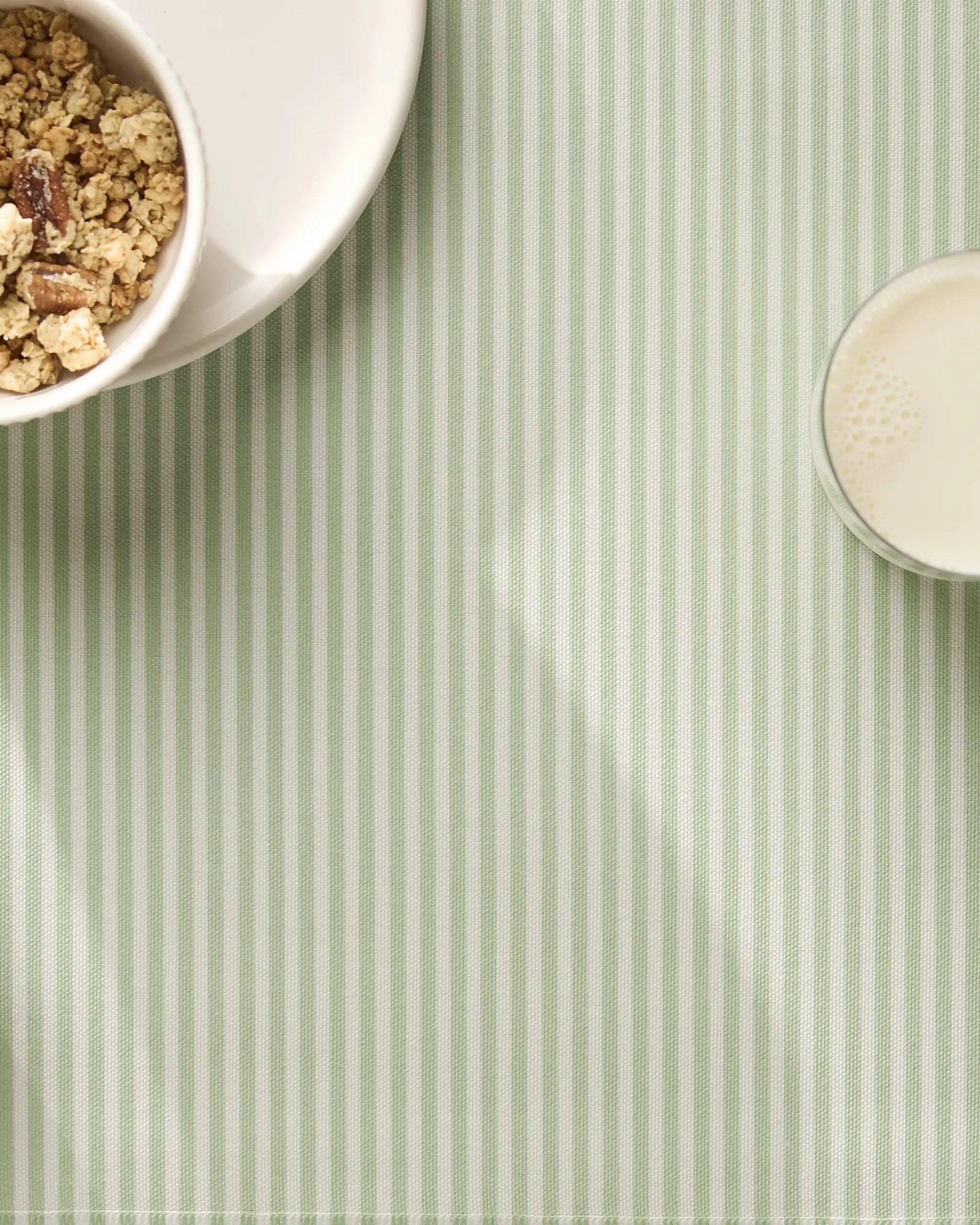 A white bowl of granola and a glass of milk rest on durable, machine-washable Geometry Seagrass placemats, both partially visible in the top corners on a pale green and white striped tablecloth.