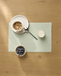 A bowl of granola, a glass of milk, and blueberries are arranged on Geometry brand Seagrass placemats with light green stripes on a wooden table.