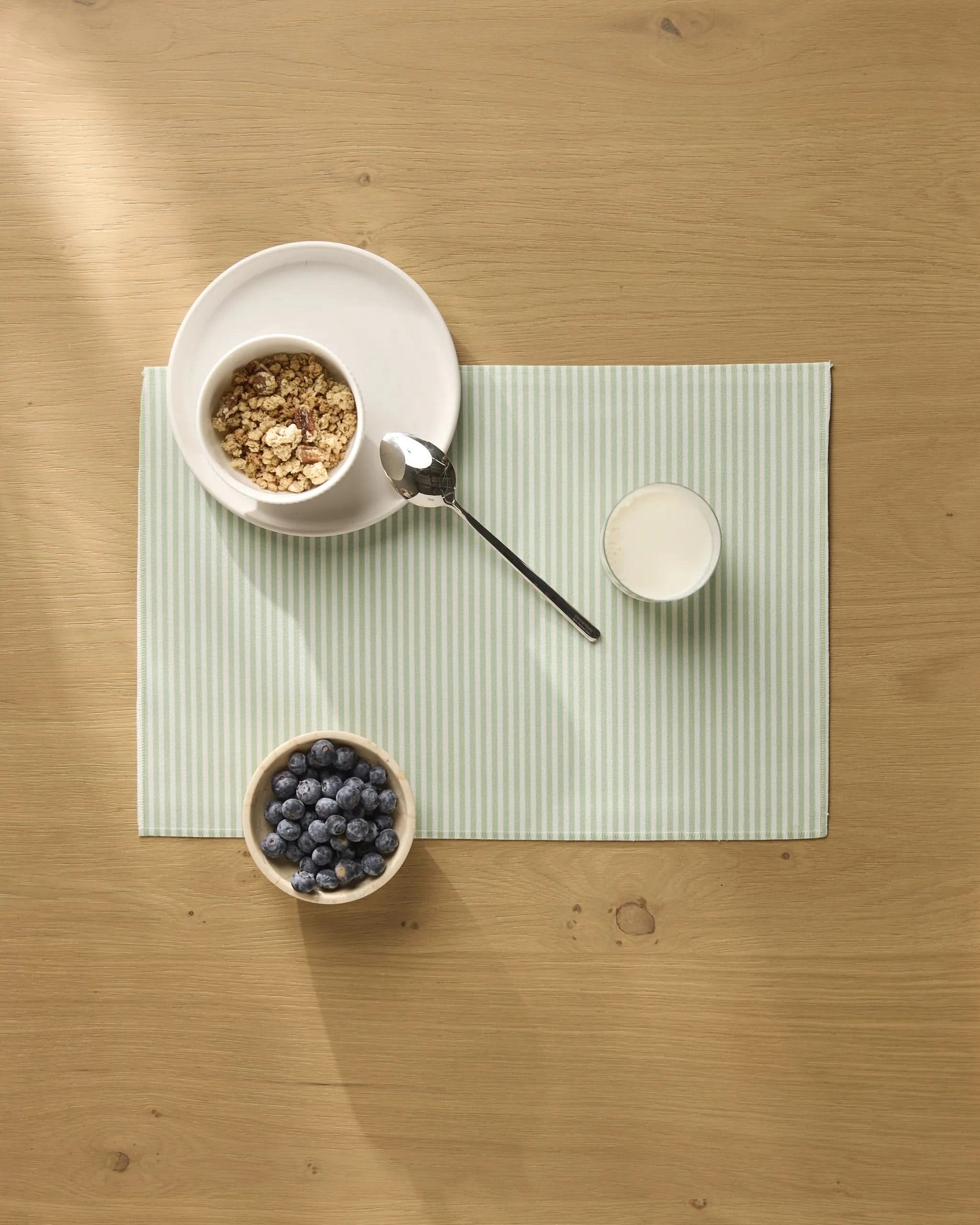 A bowl of granola, a glass of milk, and blueberries are arranged on Geometry brand Seagrass placemats with light green stripes on a wooden table.