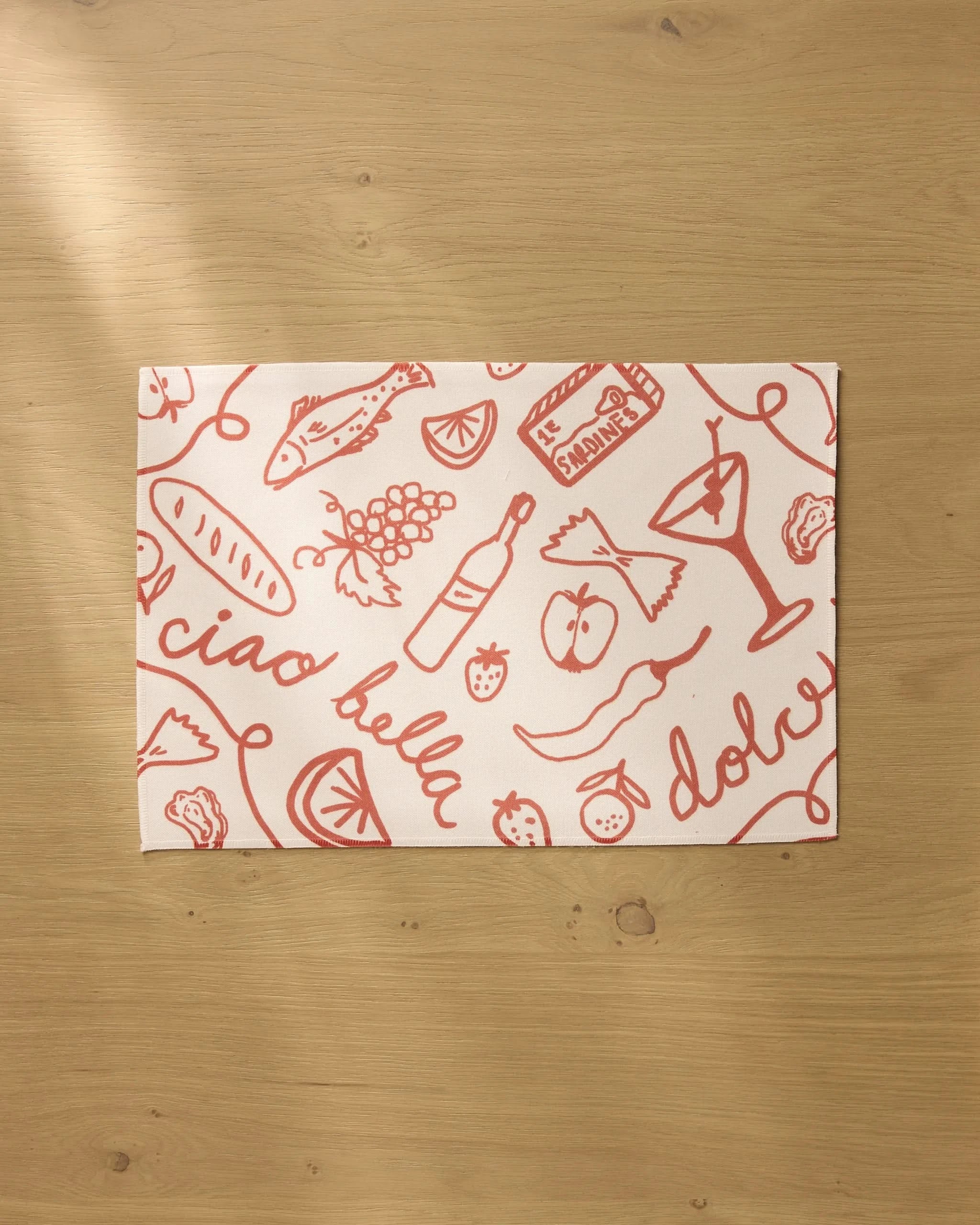 The Summer Air placemat by Geometry features red Italian-themed sketches and phrases on white recycled fabric, resting on a light wooden surface. Both stylish and practical, these placemats are machine washable.
