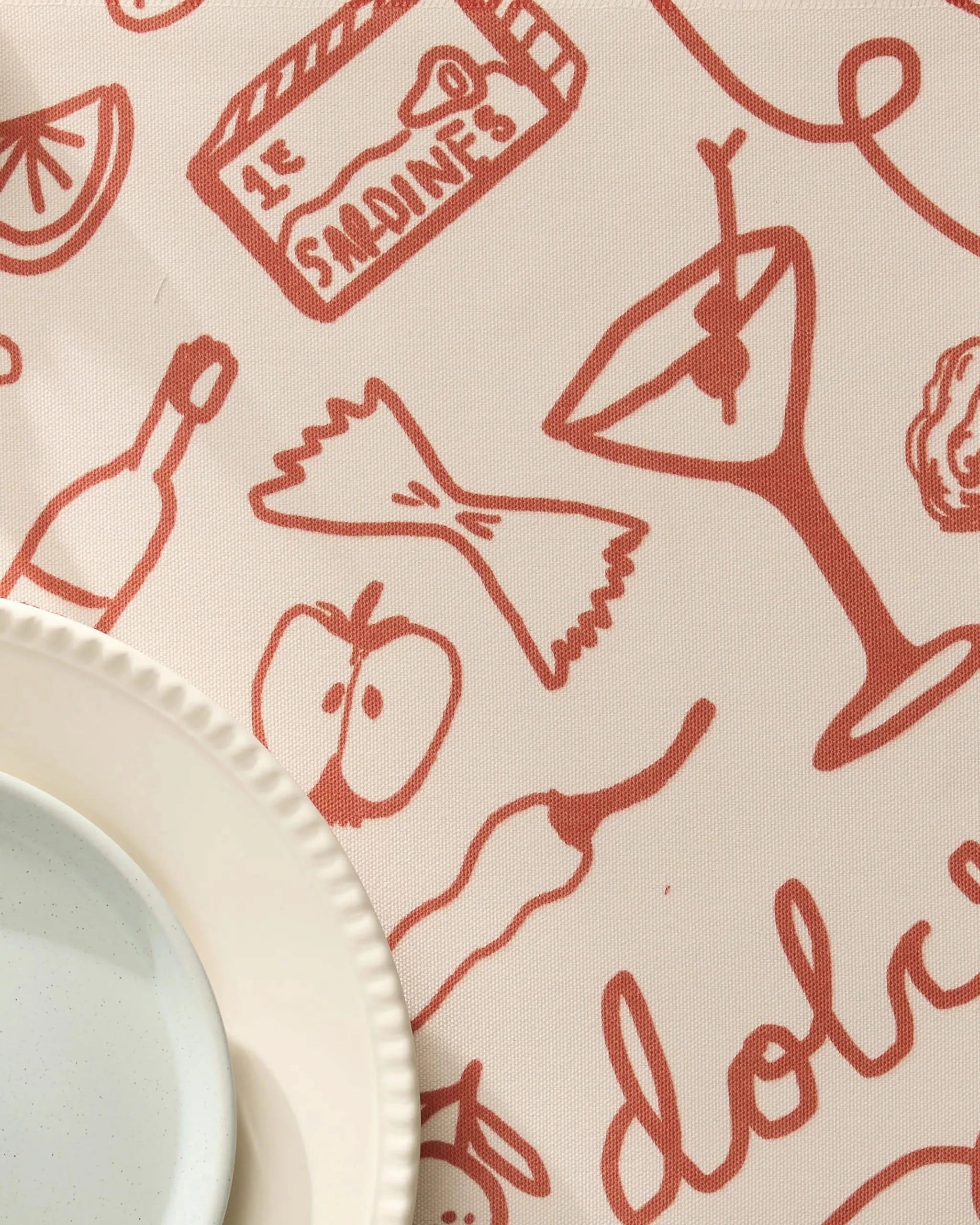 A close-up of Geometrys Summer Air tablecloth, featuring red food-themed illustrations. A white plate rests on recycled placemats in the bottom left corner.