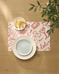 A table setting featuring Geometry’s Summer Air placemats with red food-themed illustrations, a white plate, pale blue bowl, and apple slices in a small dish sits beside a leafy green plant. All placemats are machine washable.
