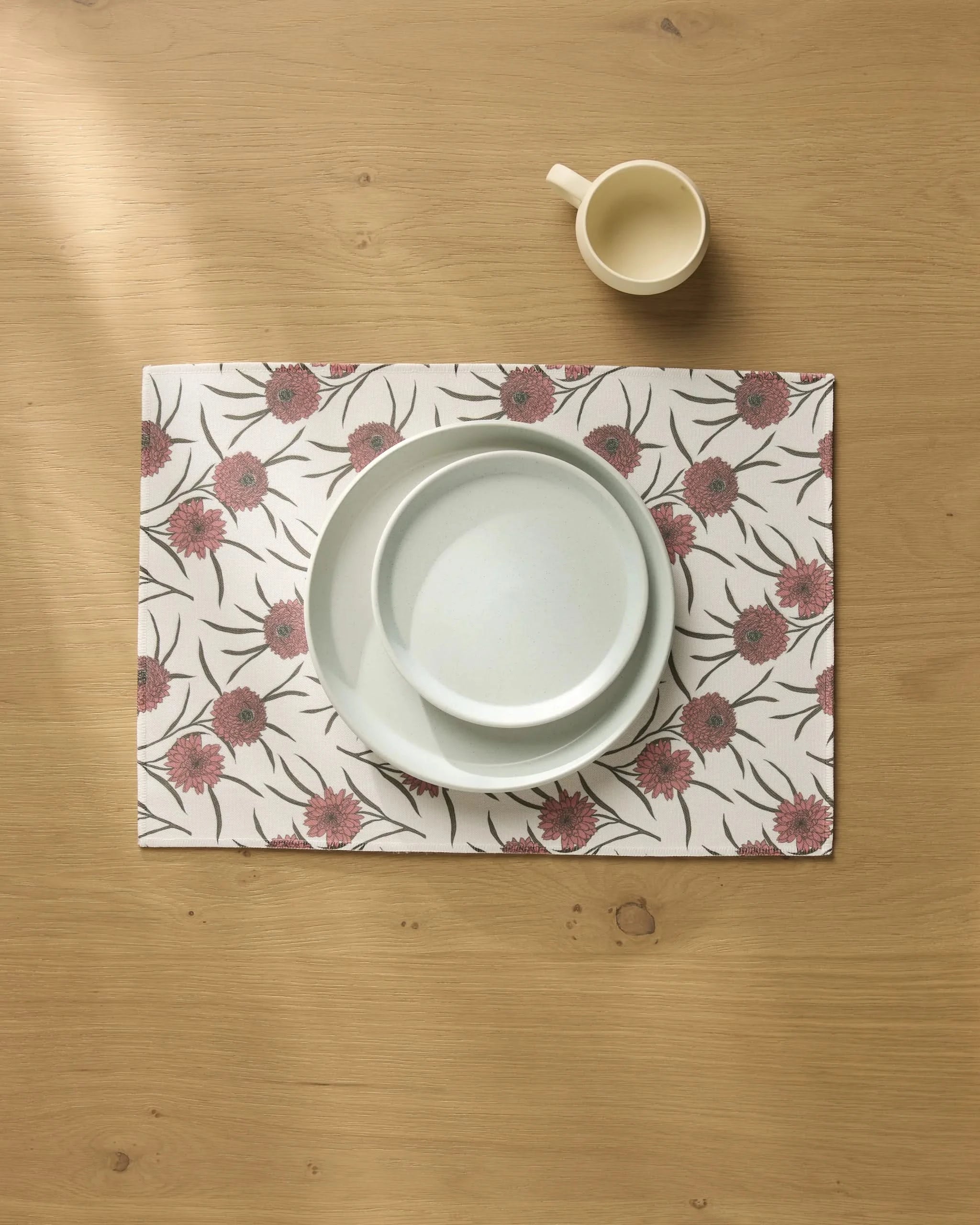 A white mug and two white plates are stacked on the Geometry Wild Dahlia recycled placemat, featuring red and purple flowers, all arranged on a light wood table.