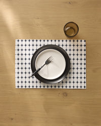 The Geometry Espresso Mosaic placemat holds a black and white plate with a fork on a wooden table, while an empty brown-tinted glass sits in the top right corner.