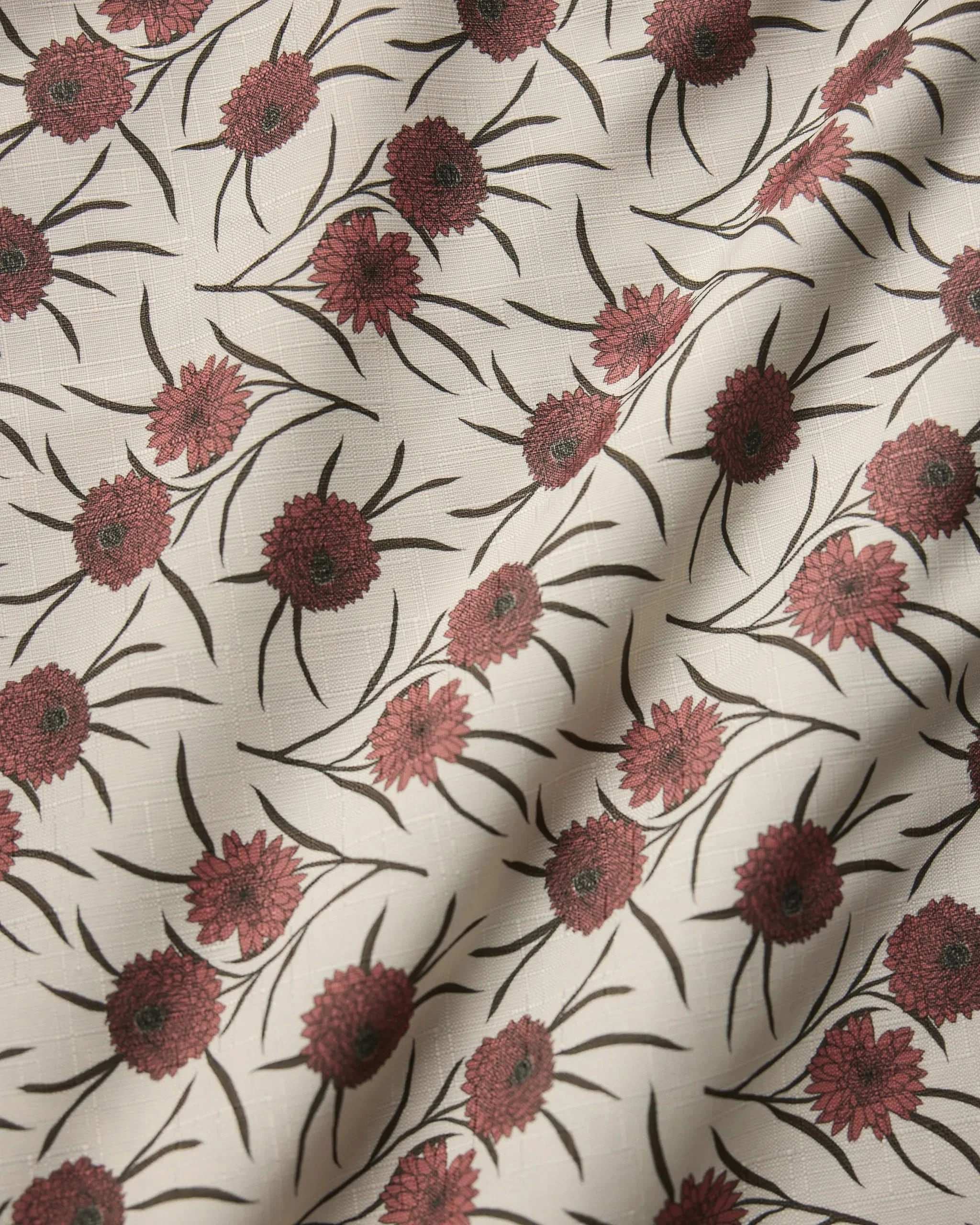 The Wild Dahlia tablecloth by Geometry is cream-colored and stain-resistant, featuring a repeating pattern of red flowers with green stems, accented by subtle folds and soft shadows.