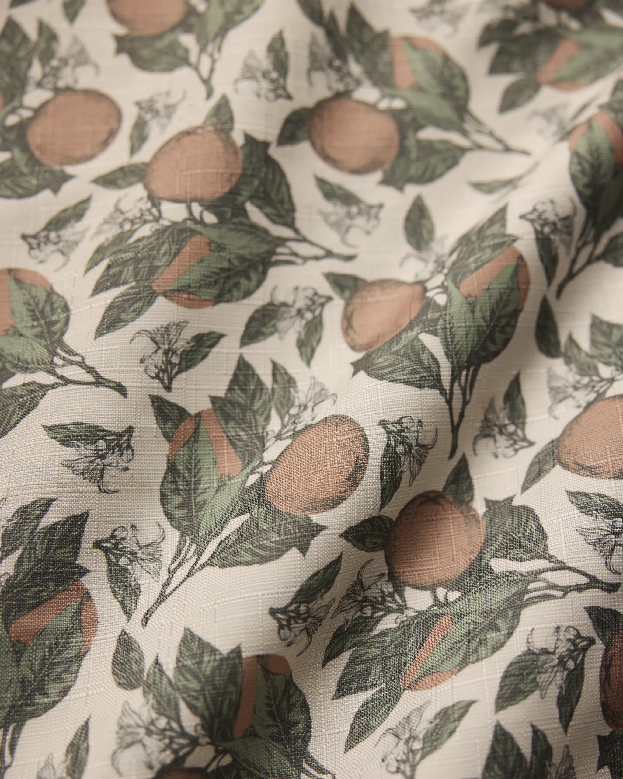 A close-up of Geometrys Valencia table runner shows its stain-resistant fabric with a botanical print of oranges, green leaves, and white blossoms on a light background. The texture and weave are clearly visible.