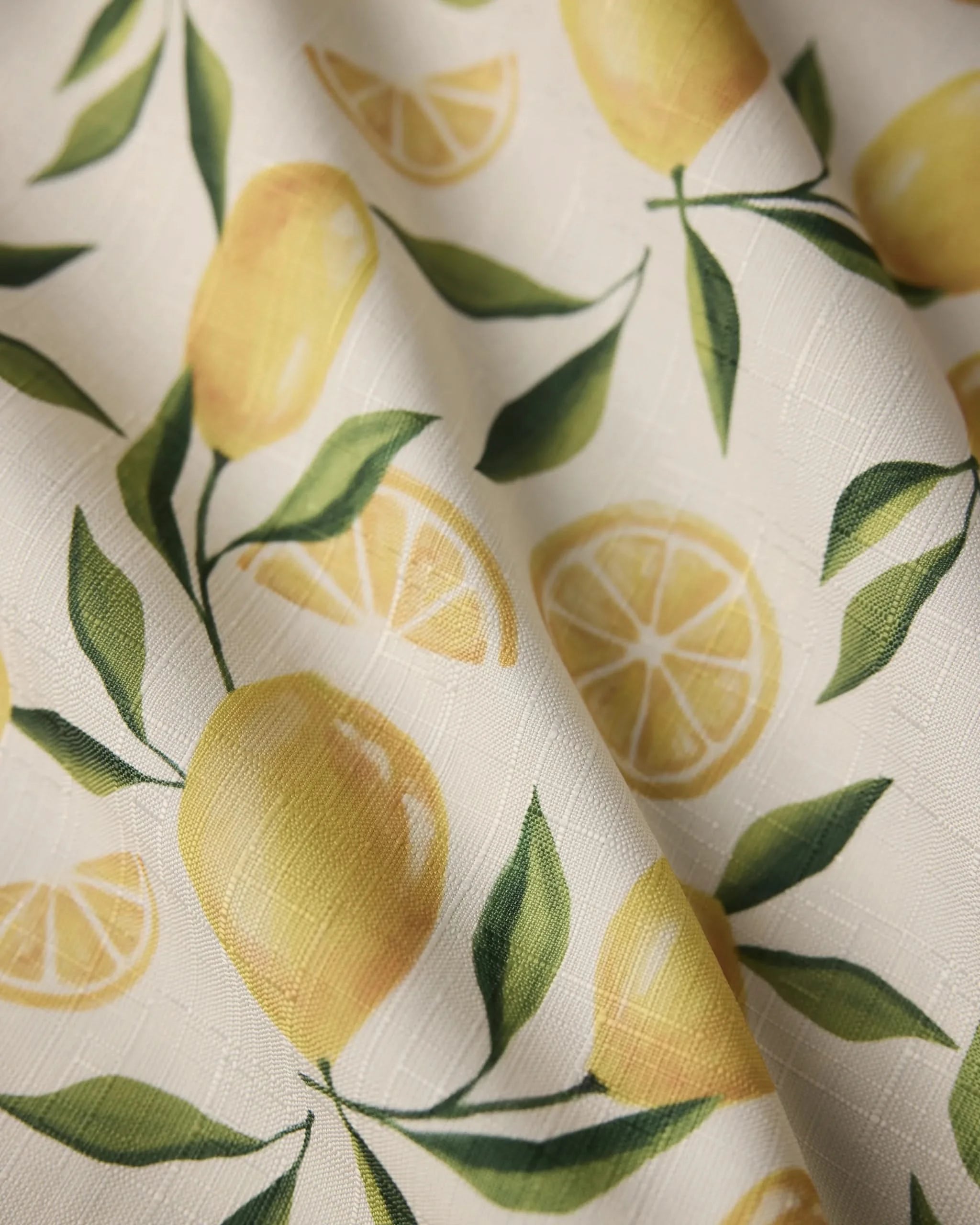 A close-up of Geometrys Limoncello recycled table runner, featuring a printed pattern of whole lemons, lemon slices, and green leaves on a subtly textured light background.