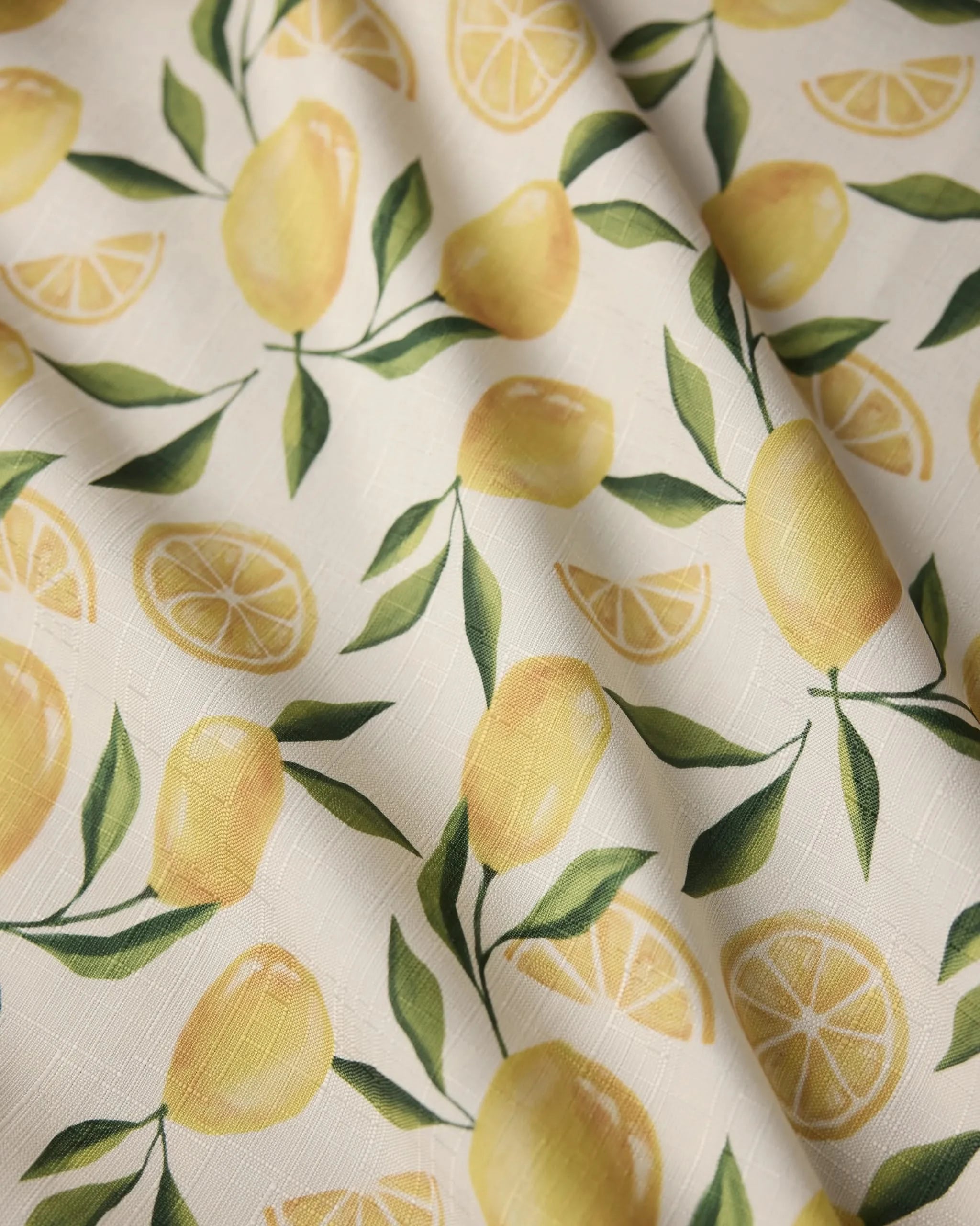 A close-up of Geometry’s Limoncello tablecloth shows a stain-resistant fabric with whole lemons, slices, and green leaves on a white textured background. Some wrinkles are visible.