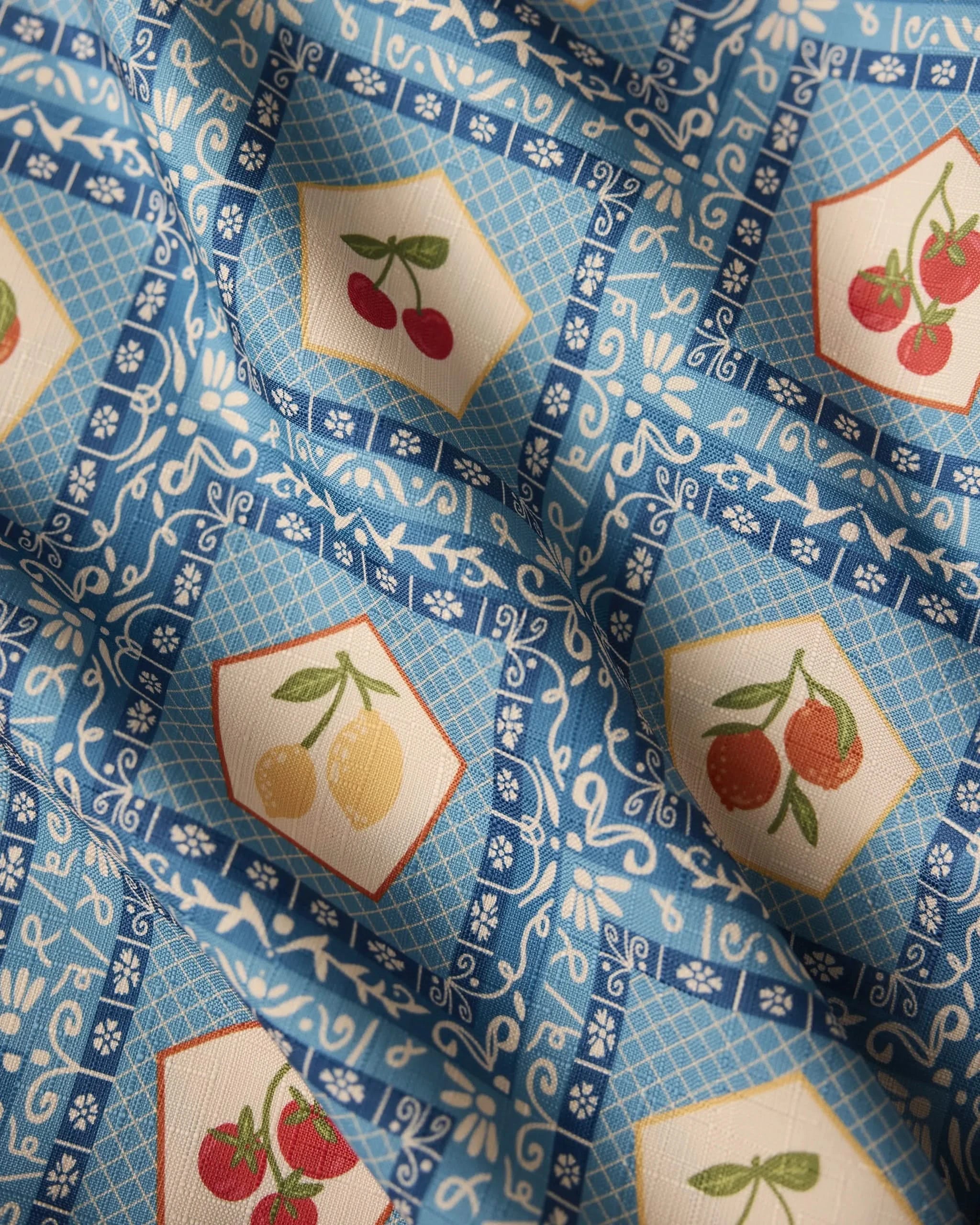 The Geometry Italian Tiles tablecloth features a blue and white pattern with hexagonal frames showcasing cherries, lemons, and tomatoes. Its lightweight, slightly wrinkled fabric adds texture and subtle shadowing to the surface.