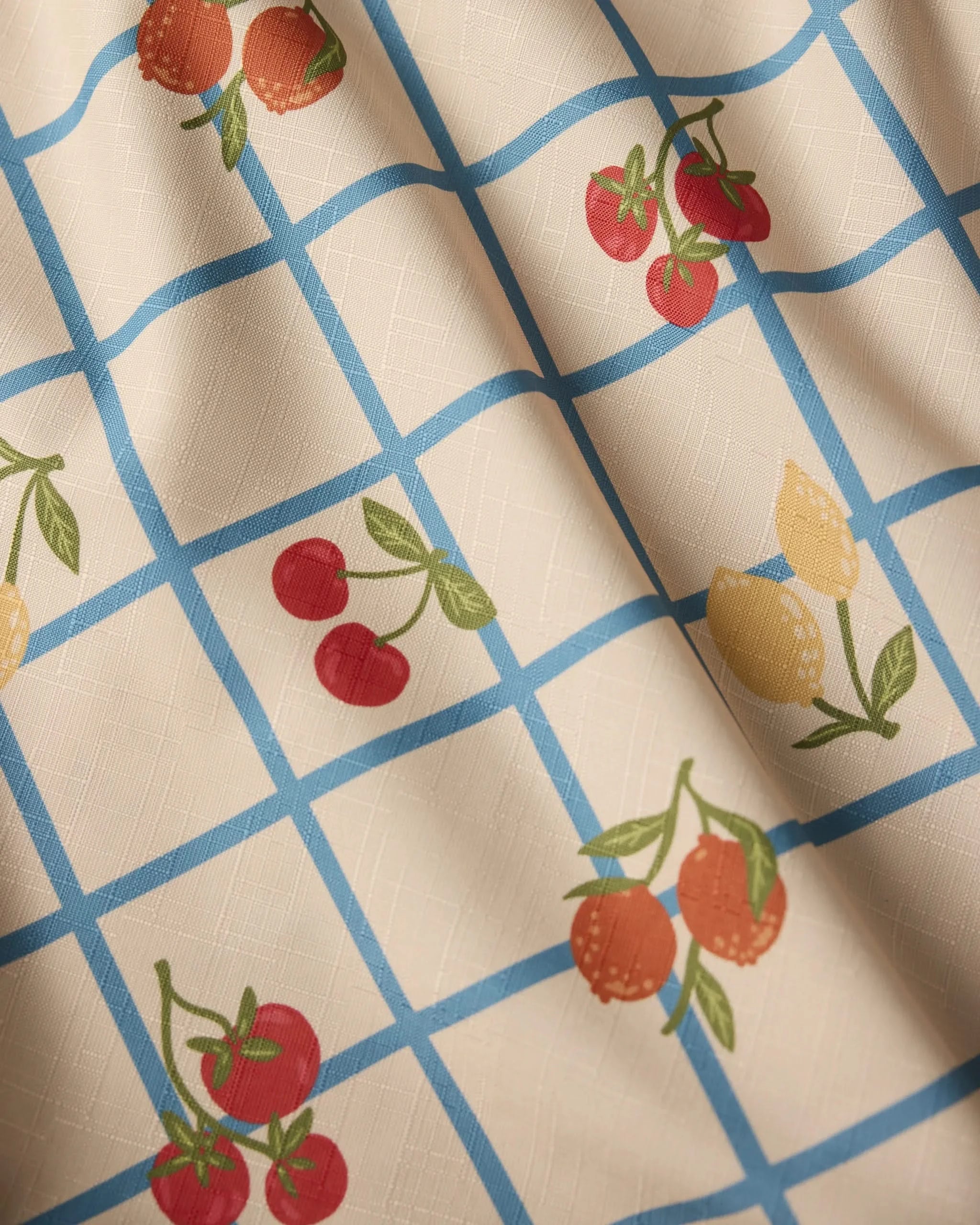 Geometry’s Italian Summer Grid tablecloth is lightweight with blue grid lines on a cream background, featuring cherries, lemons, and oranges in each square. The fabric shows subtle folds and soft shadows.
