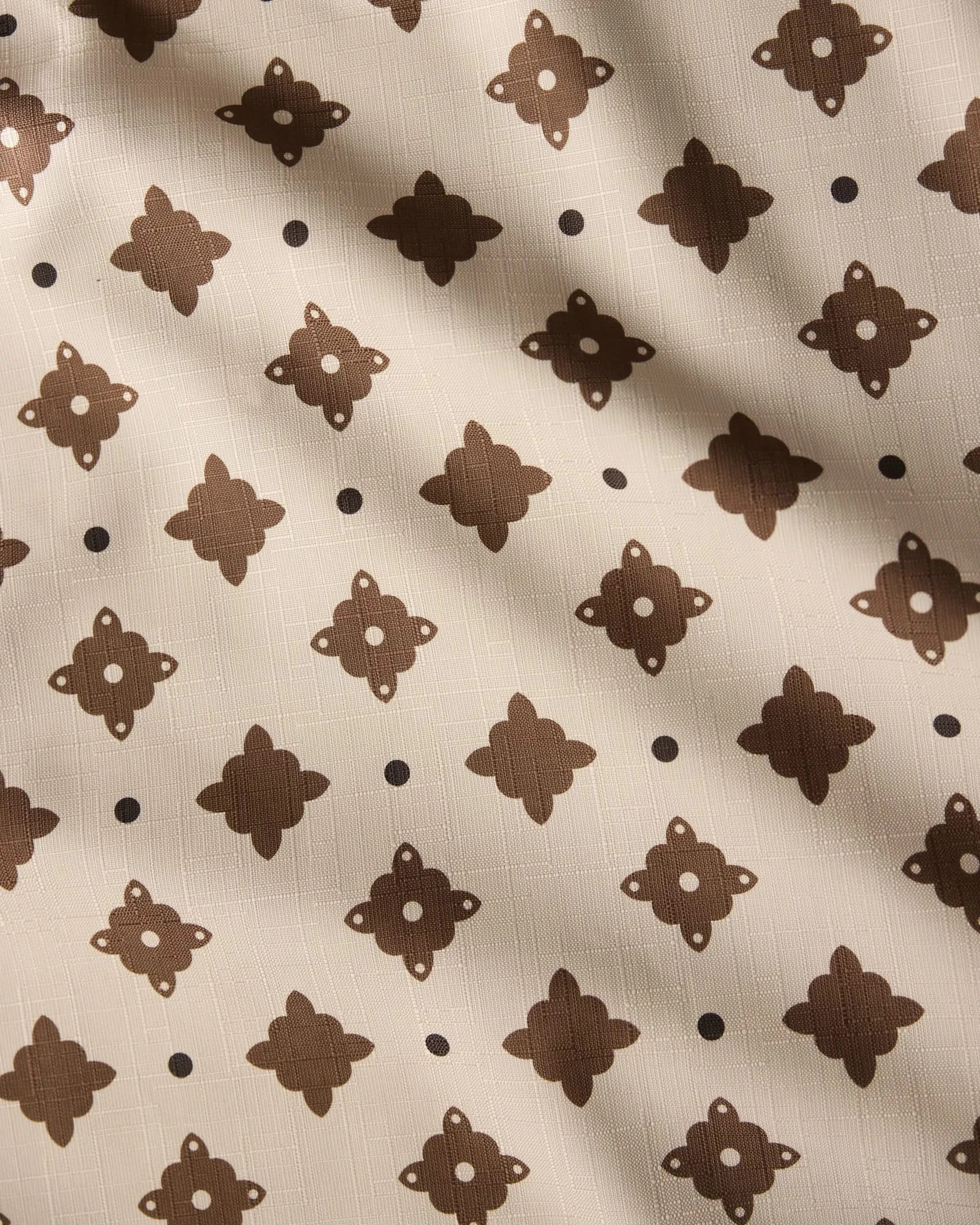 The Heritage Stamp tablecloth by Geometry features beige fabric with a repeating dark brown geometric pattern and dots, accented by subtle diagonal shadows—an eco-friendly choice perfect for modern homes.