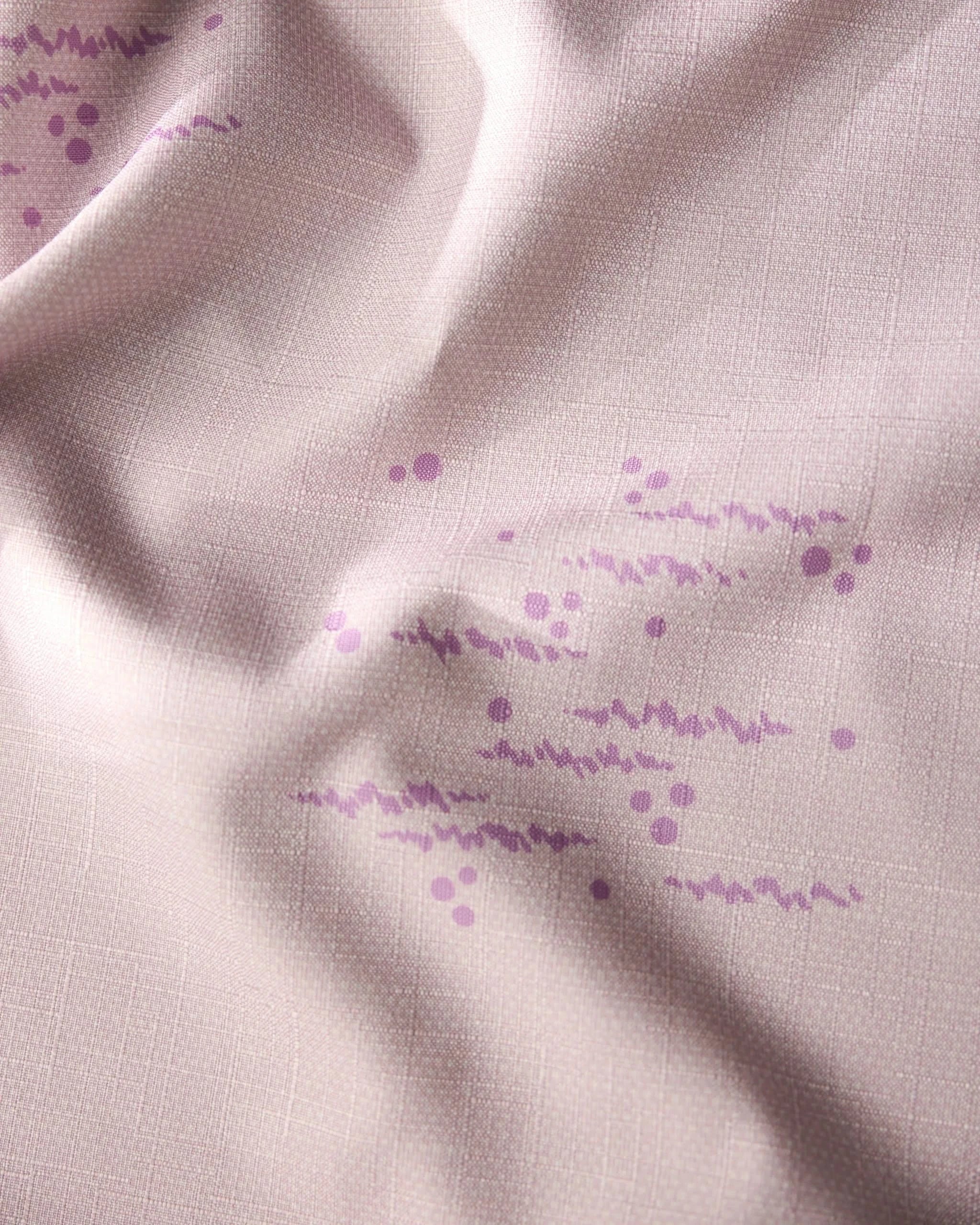 A close-up of Geometrys Bubble Ditzy Dusty Rose tablecloth shows stain-resistant light pink fabric with a subtle grid, soft texture, and gentle folds, decorated with clusters of small purple dots and wavy lines.