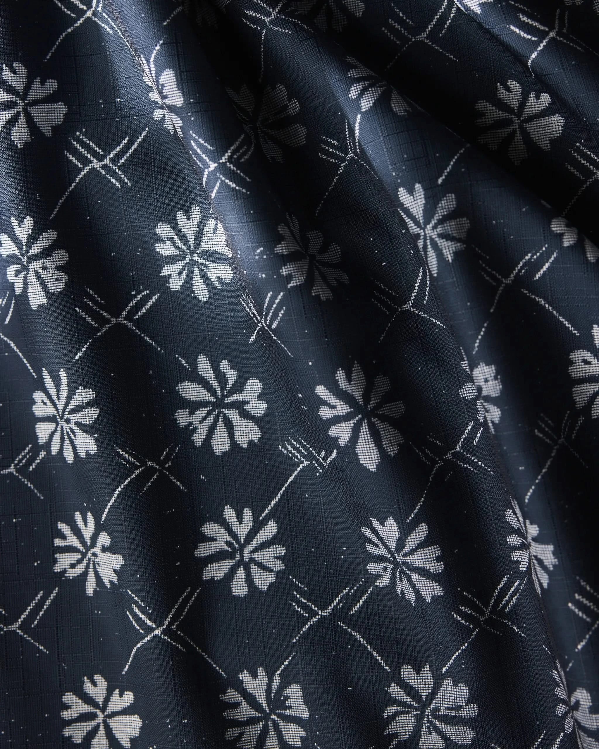 Bonnie Indigo by Geometry is a dark blue, stain-resistant tablecloth with a repeating white floral pattern of stylized flowers and intersecting diagonal lines. Its slightly textured fabric drapes elegantly, creating soft folds and shadows.