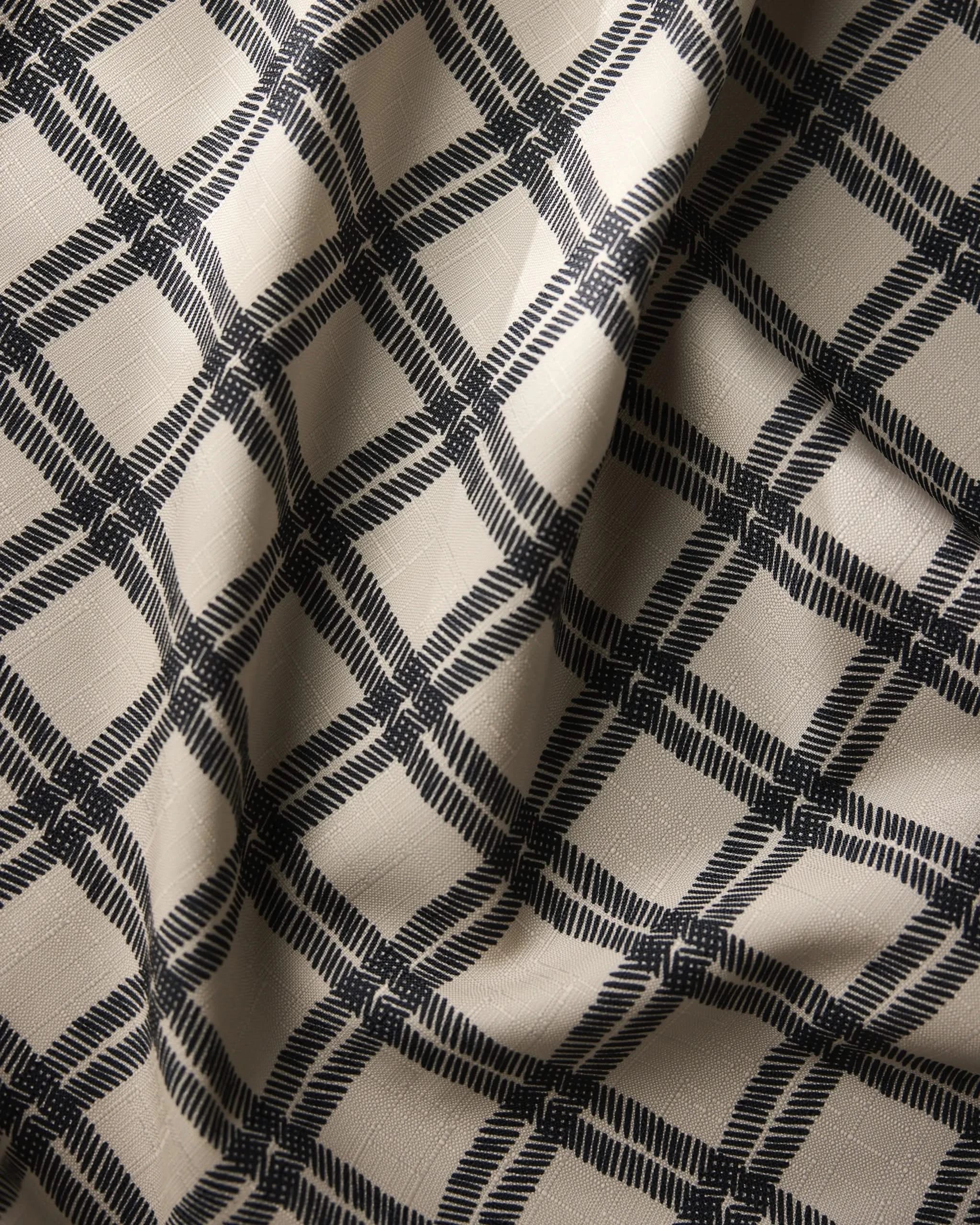 A close-up of Geometrys Blackstone tablecloth shows its beige, stain-resistant fabric with a black plaid pattern, highlighted by soft folds and shadows that add texture and depth.