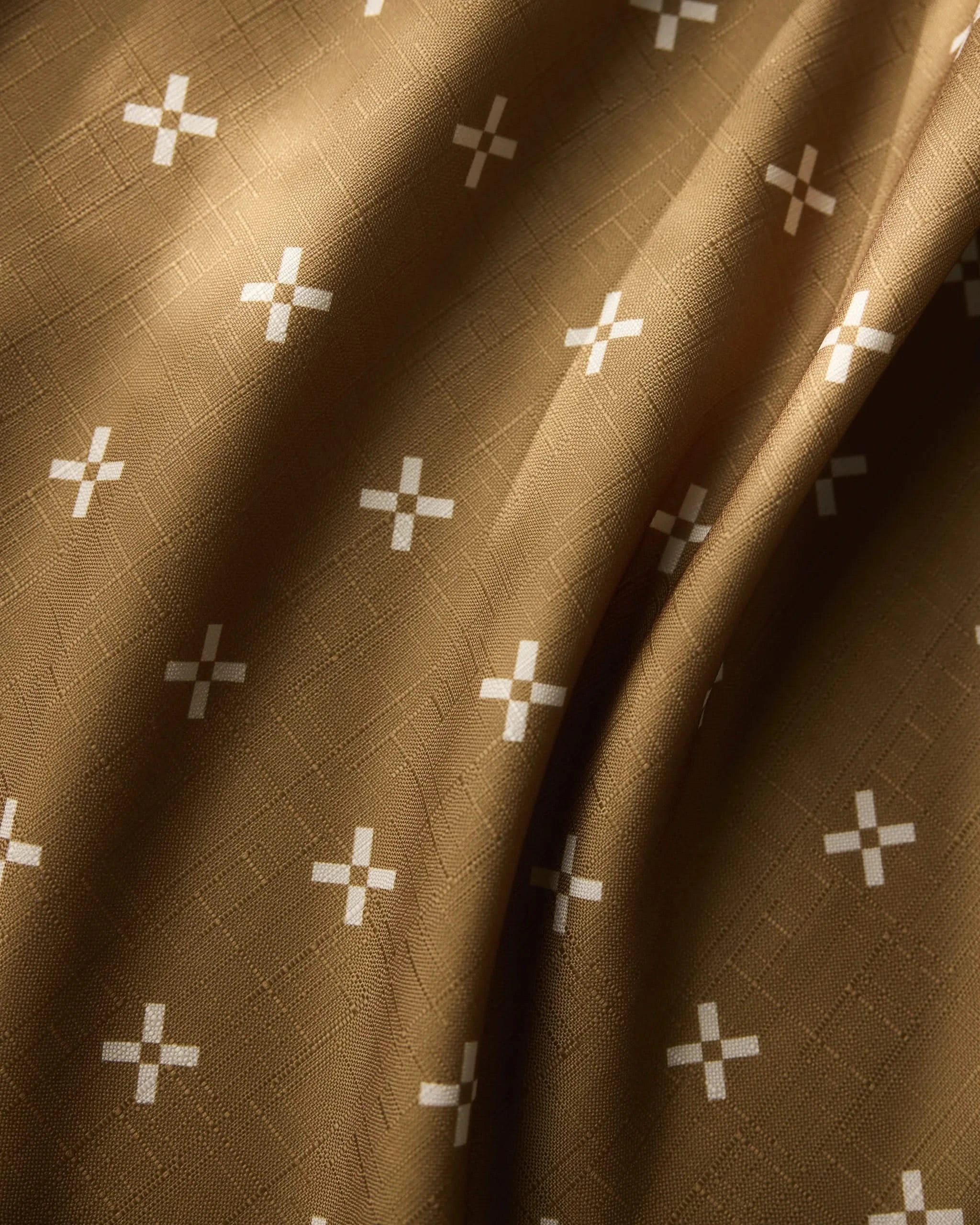 Amber Stitch by Geometry is an eco-friendly tablecloth made of brown fabric with a subtle texture and evenly spaced white plus-sign designs—shown with gentle folds and soft, natural lighting, it’s perfect for contemporary spaces.