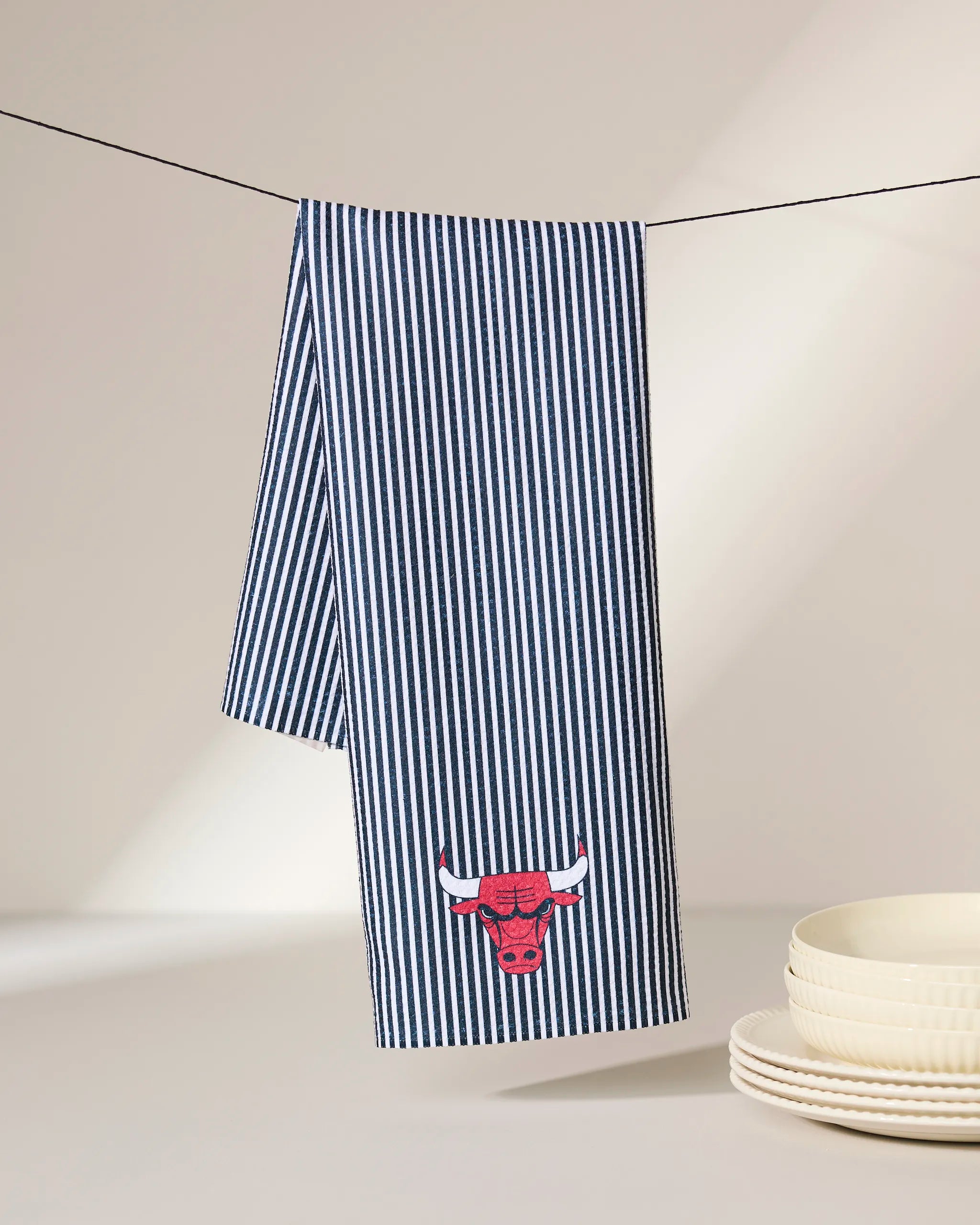 A Bulls Classic tea towel by Geometry, featuring blue and white stripes with a red Chicago Bulls logo, hangs on a clothesline. Its ultra-absorbent fabric awaits use next to a stack of white dishes on a neutral background.
