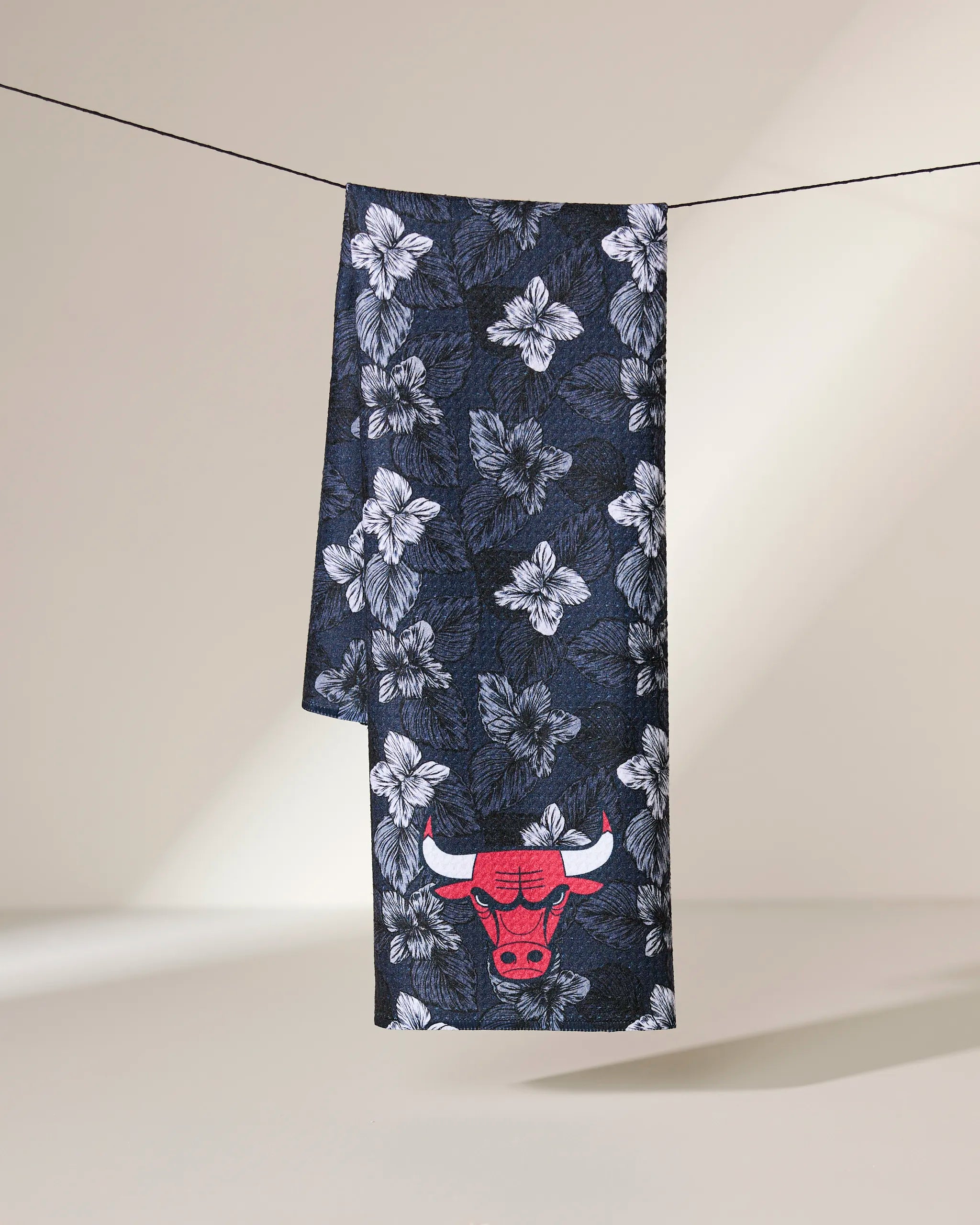 The Bulls Floral tea towel by Geometry features a dark blue and white floral design with a red Chicago Bulls logo, hanging over a black clothesline against a neutral background—ideal for showcasing team spirit in your kitchen.
