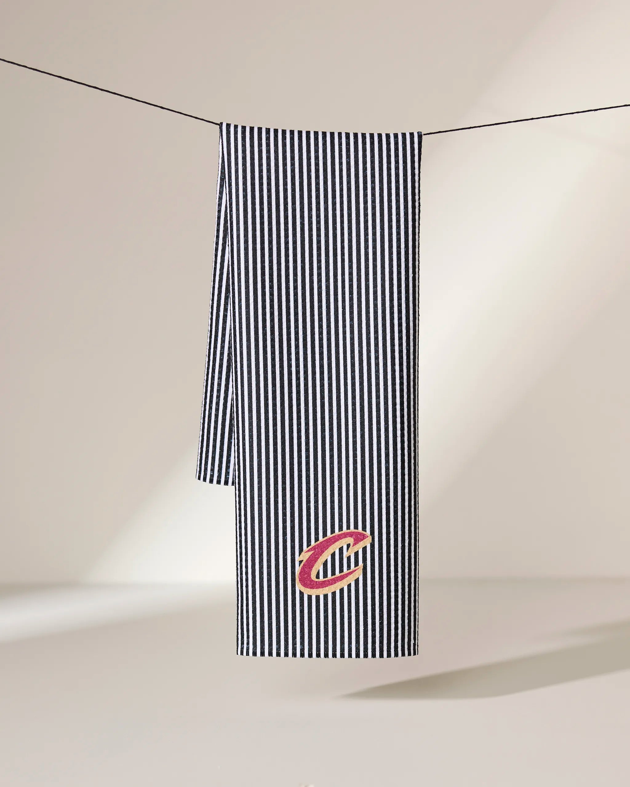 The Cavaliers Classic tea towel by Geometry hangs from a line, featuring black and white vertical stripes, a red and gold C logo near the bottom edge, and is crafted from recycled materials. The softly lit background highlights its ultra-absorbent quality.