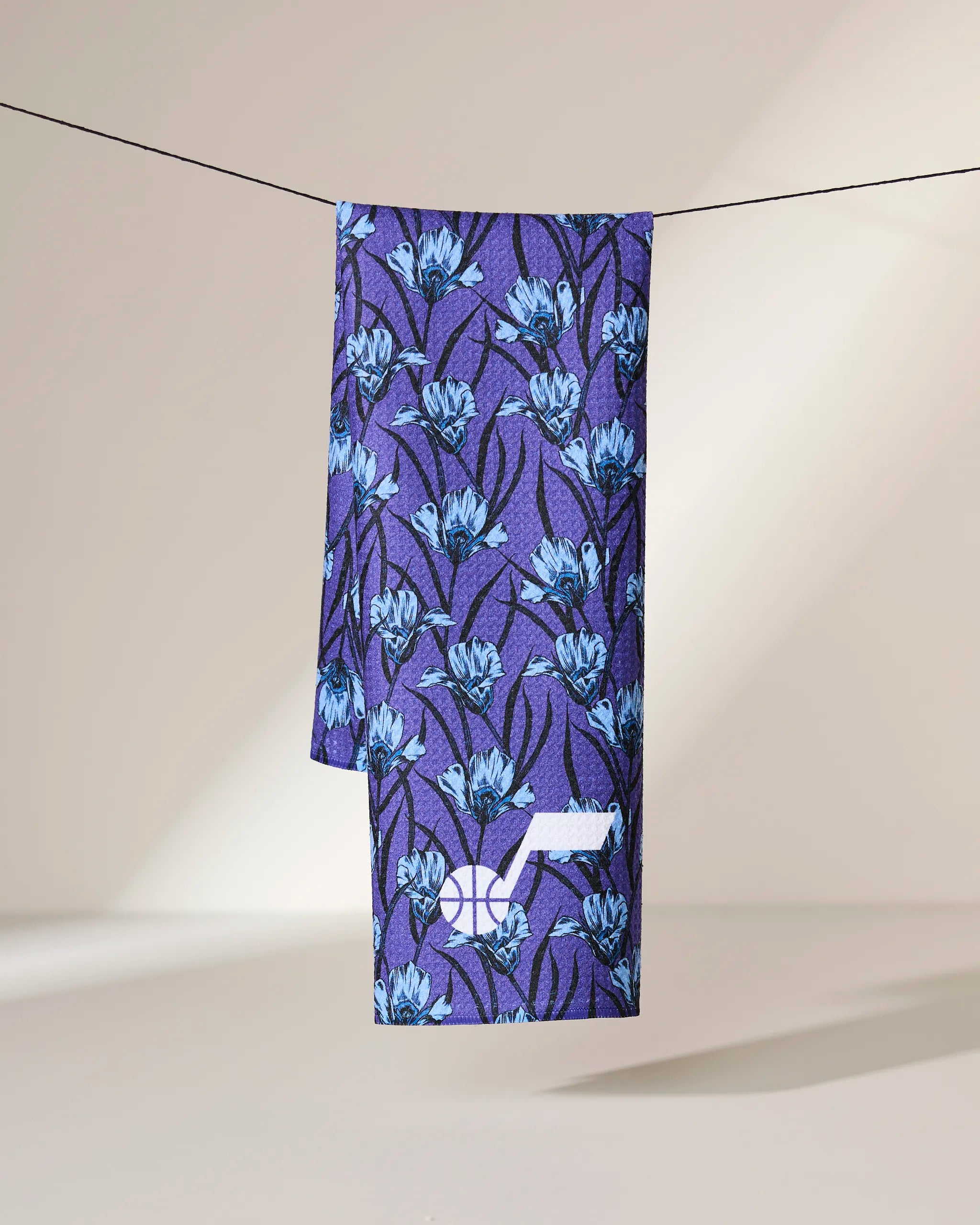 The Geometry Jazz Floral necktie, featuring a blue floral pattern on purple fabric, hangs neatly over a slim black string against a softly lit, plain background.