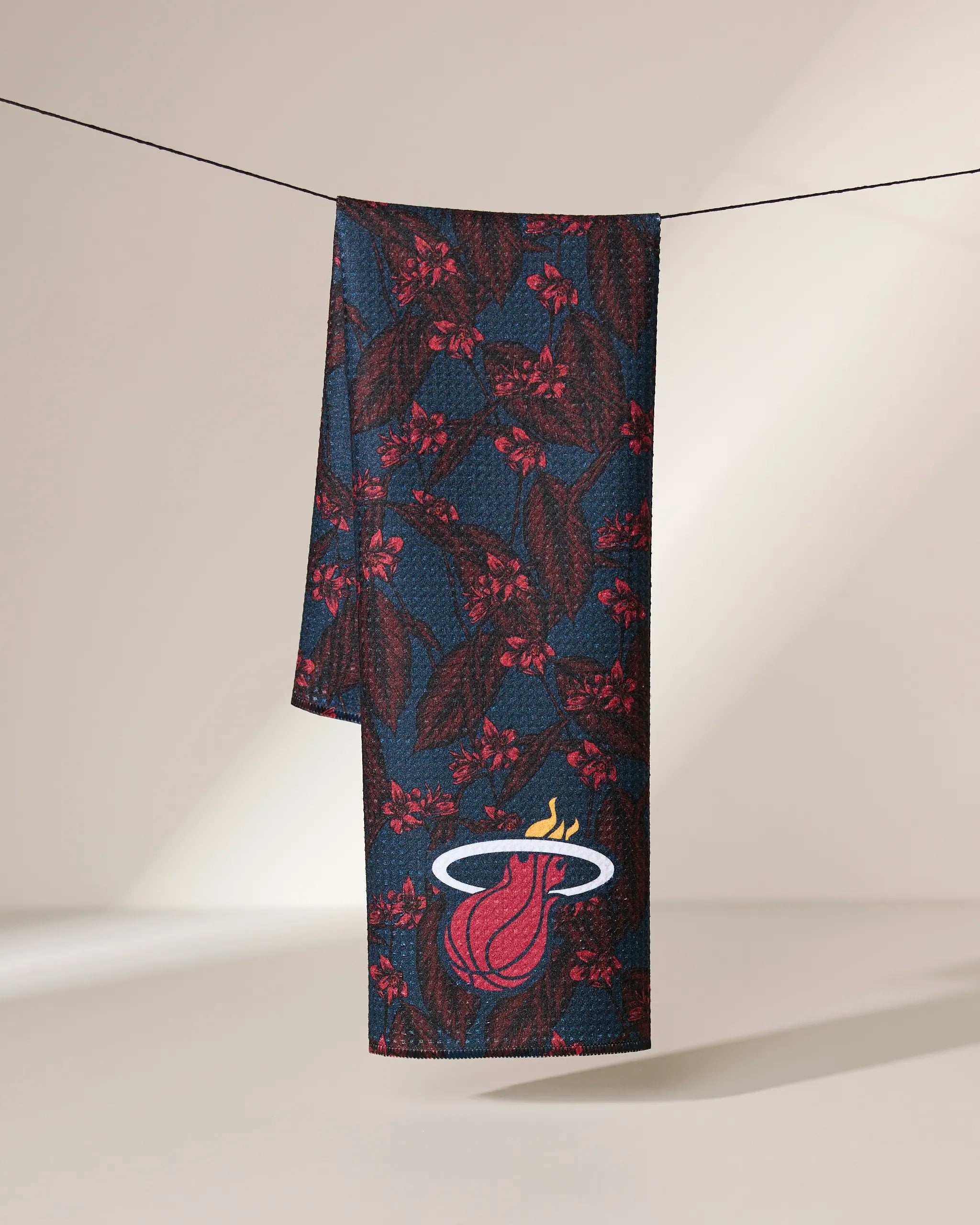 The Geometry Heat Floral towel features a red and green leafy pattern and Miami Heat logo near the bottom, hanging on a line with a softly lit neutral background.
