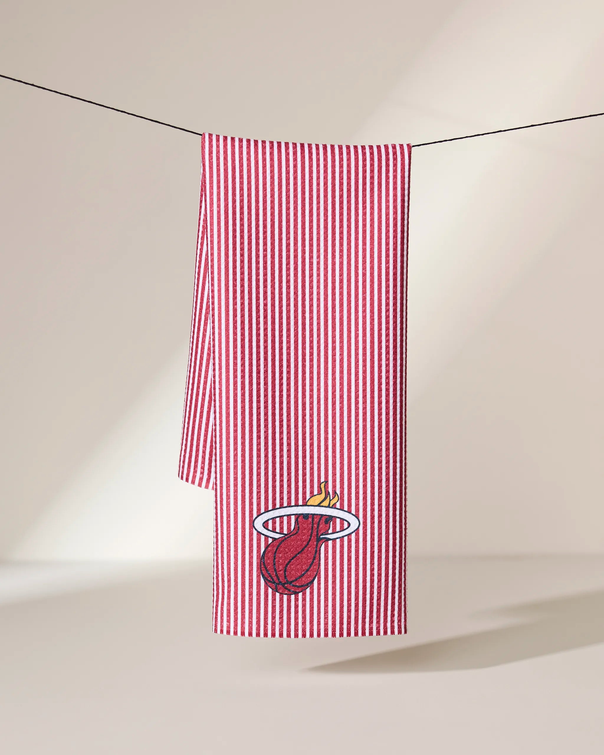 The Heat Classic kitchen towel by Geometry hangs on a line, decorated with red and white stripes and an embroidered Miami Heat flaming basketball logo near the bottom corner.