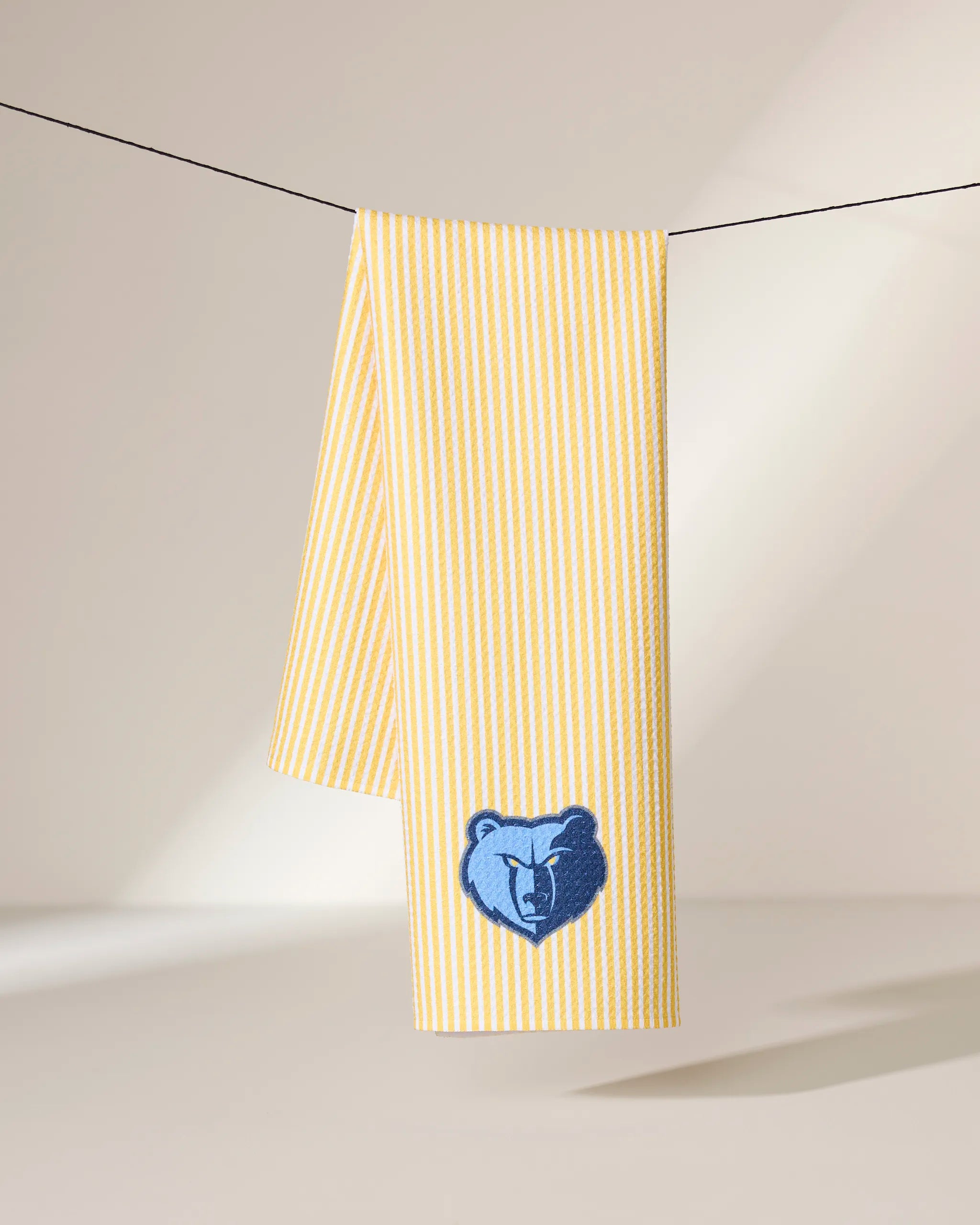 The Grizzlies Classic towel by Geometry, featuring yellow and white stripes and a blue bear head logo, hangs over a thin black line against a softly lit background.
