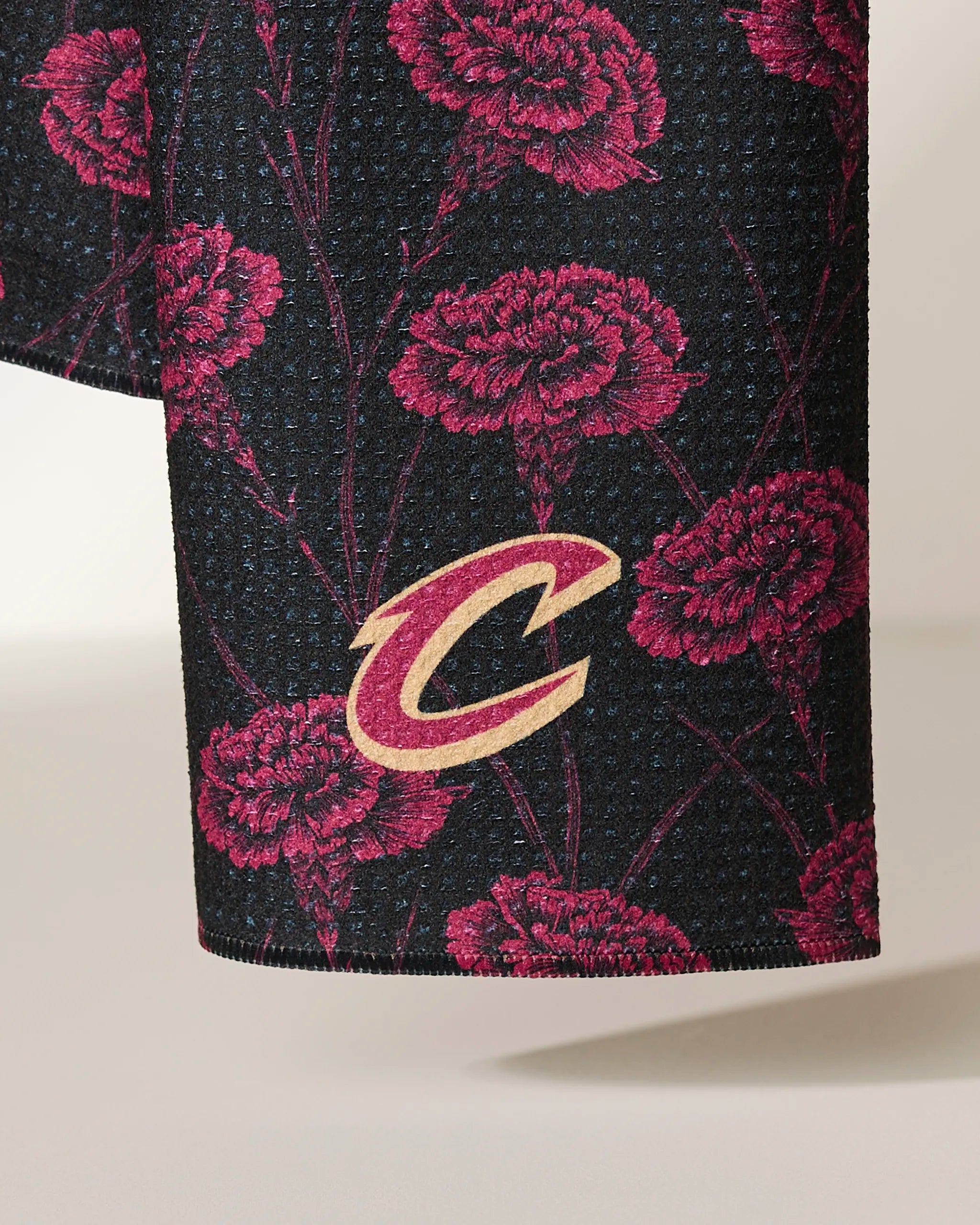 A detailed view of the Geometry Cavaliers Floral: black textured fabric from recycled materials, adorned with pink carnation patterns and a gold-and-burgundy Cleveland Cavaliers C logo.