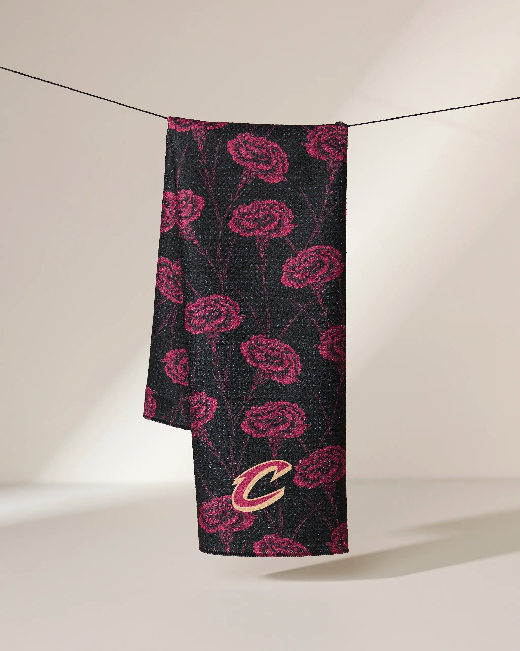 The Geometry Cavaliers Floral tea towel, featuring a pink carnation pattern and Cavaliers C logo on black, hangs over a clothesline against a neutral background.