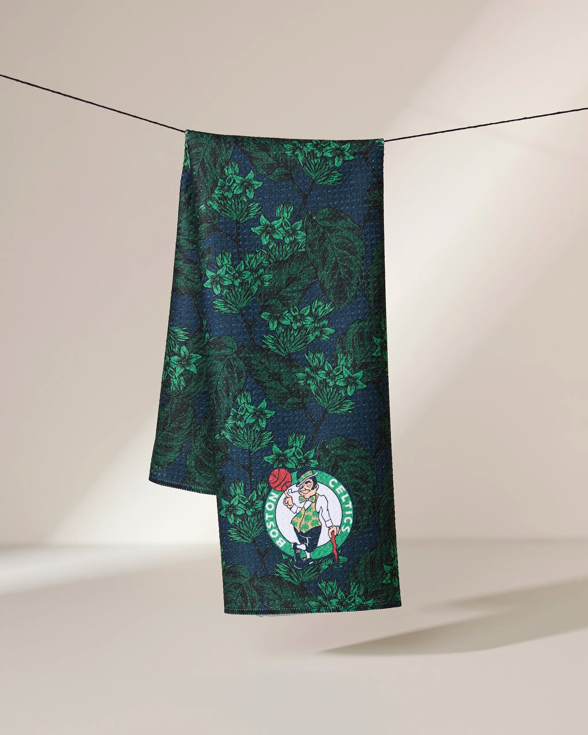 A dark green and black Celtics Floral towel by Geometry, featuring a leafy pattern and the Boston Celtics logo near the bottom corner, hangs on a line.