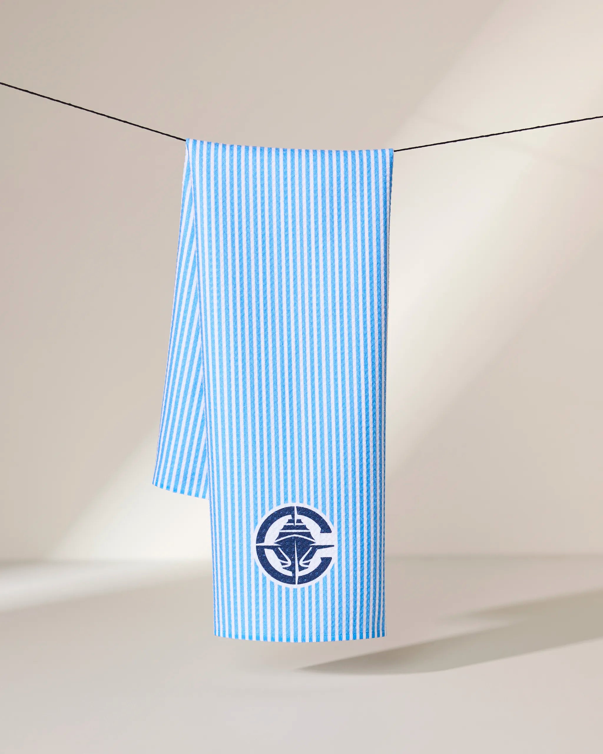 The Geometry Clippers Classic towel, featuring blue and white vertical stripes and a dark blue circular nautical emblem near the edge, hangs on a black line against a neutral background.