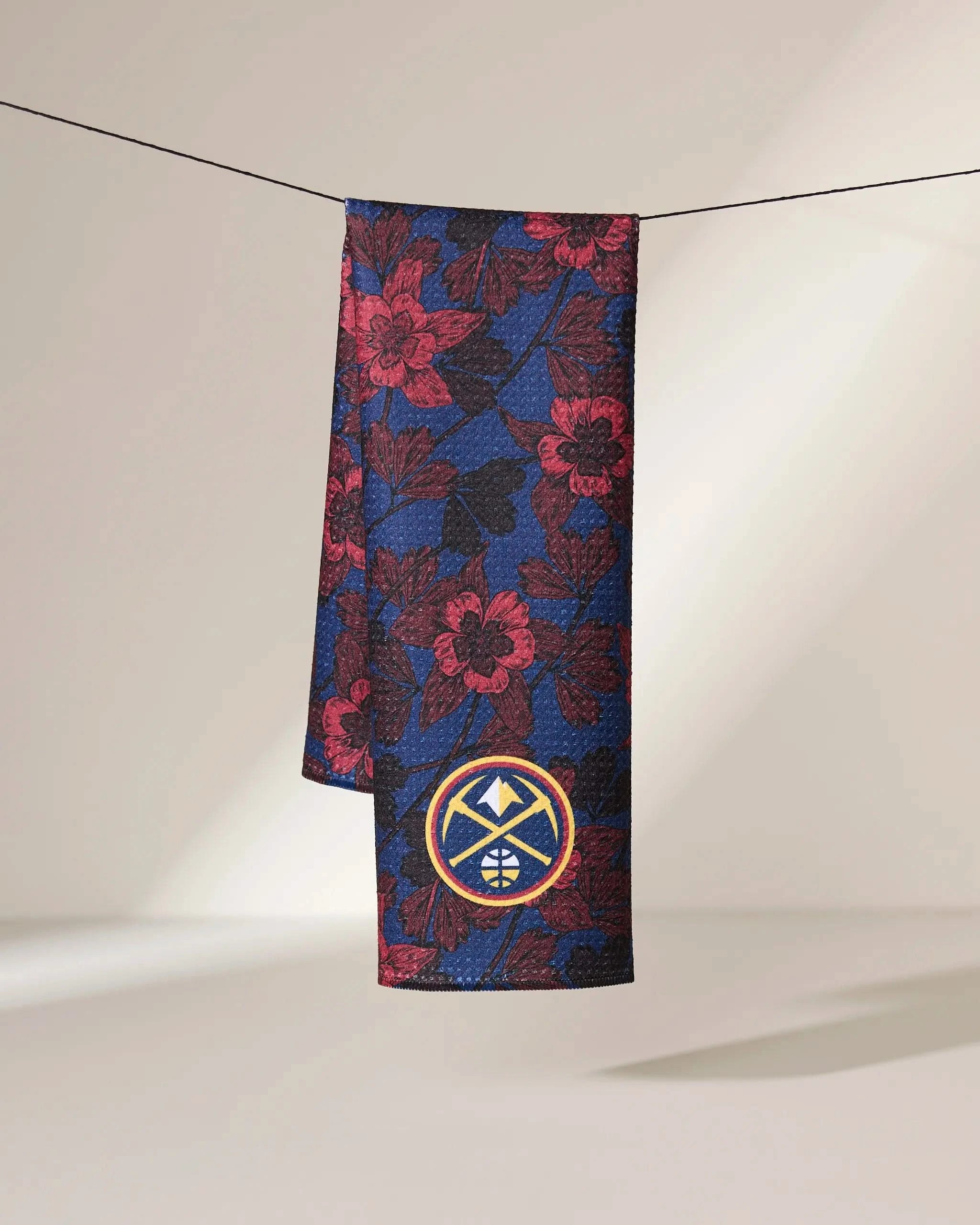 The Geometry Nuggets Floral tie, featuring a blue base with red and black floral patterns, hangs on a line. Near the bottom is an embroidered patch with a yellow border and geometric mountain-basketball design.