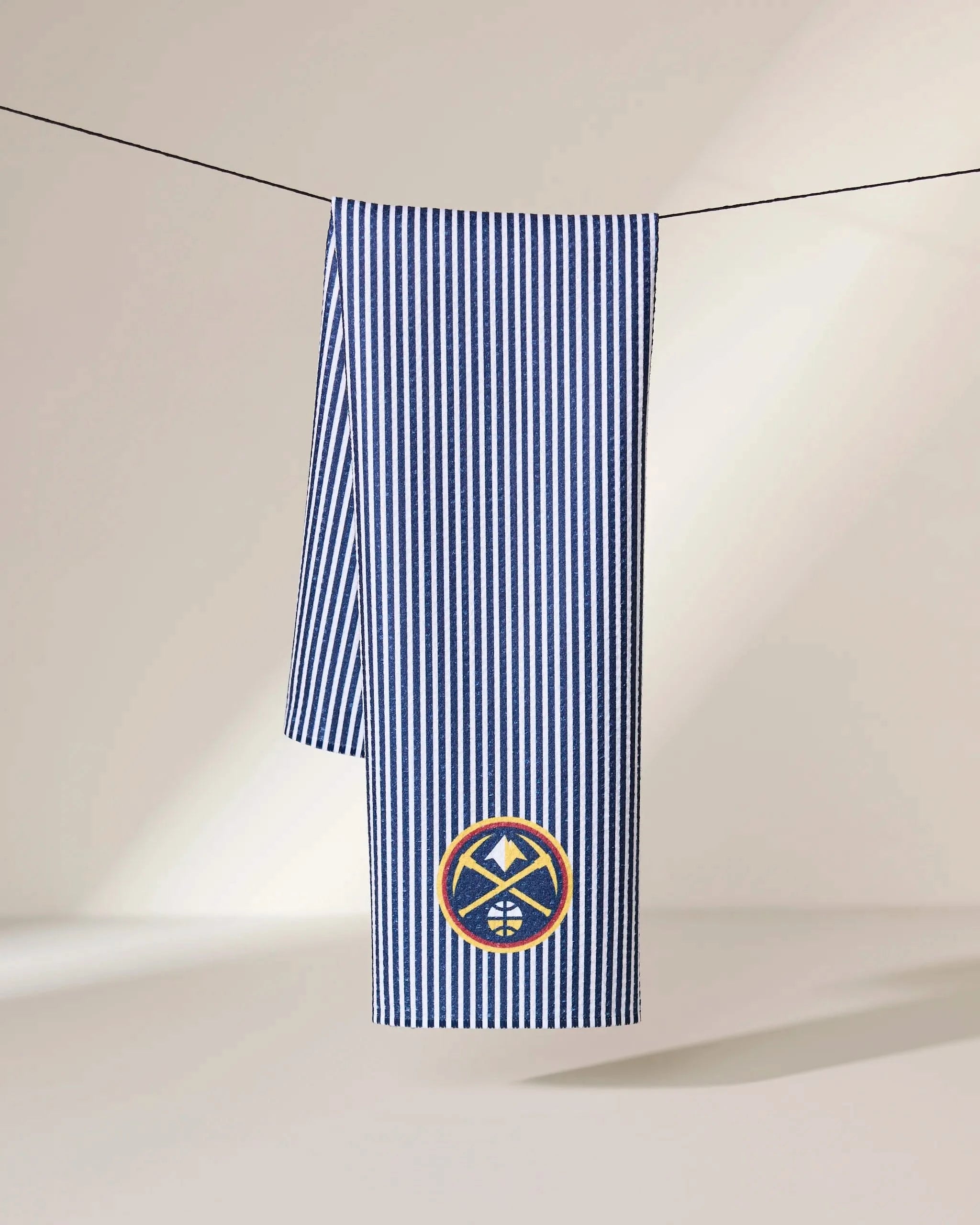 The Geometry Nuggets Classic towel in blue and white vertical stripes hangs on a black line against a light background, featuring a round patch with a mountain and basketball design near the bottom.
