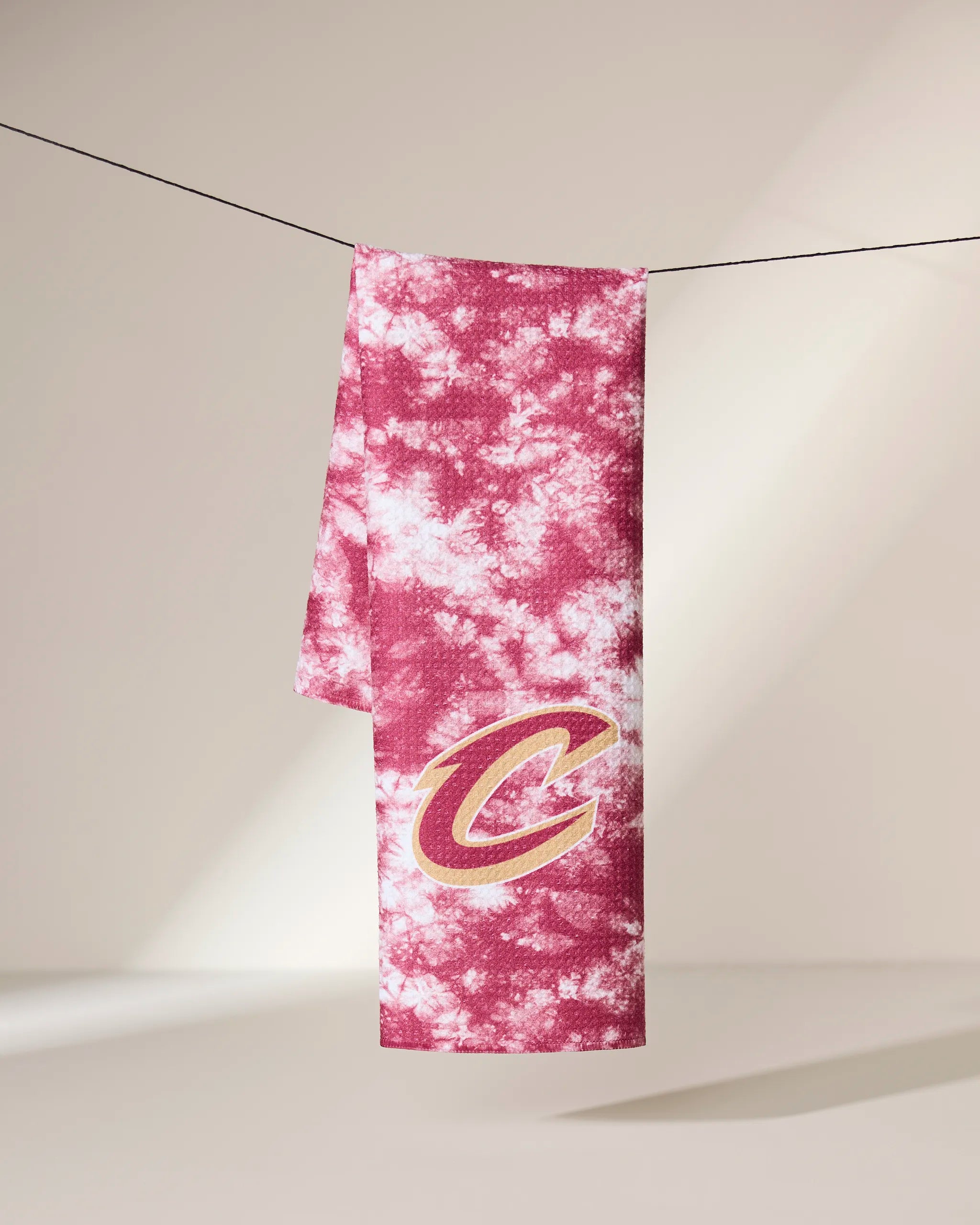 A pink and white tie-dye Cavaliers Flash tea towel by Geometry, made from quick-drying recycled materials and featuring a bold Cleveland Cavaliers C logo, hangs on a black line against a neutral background.