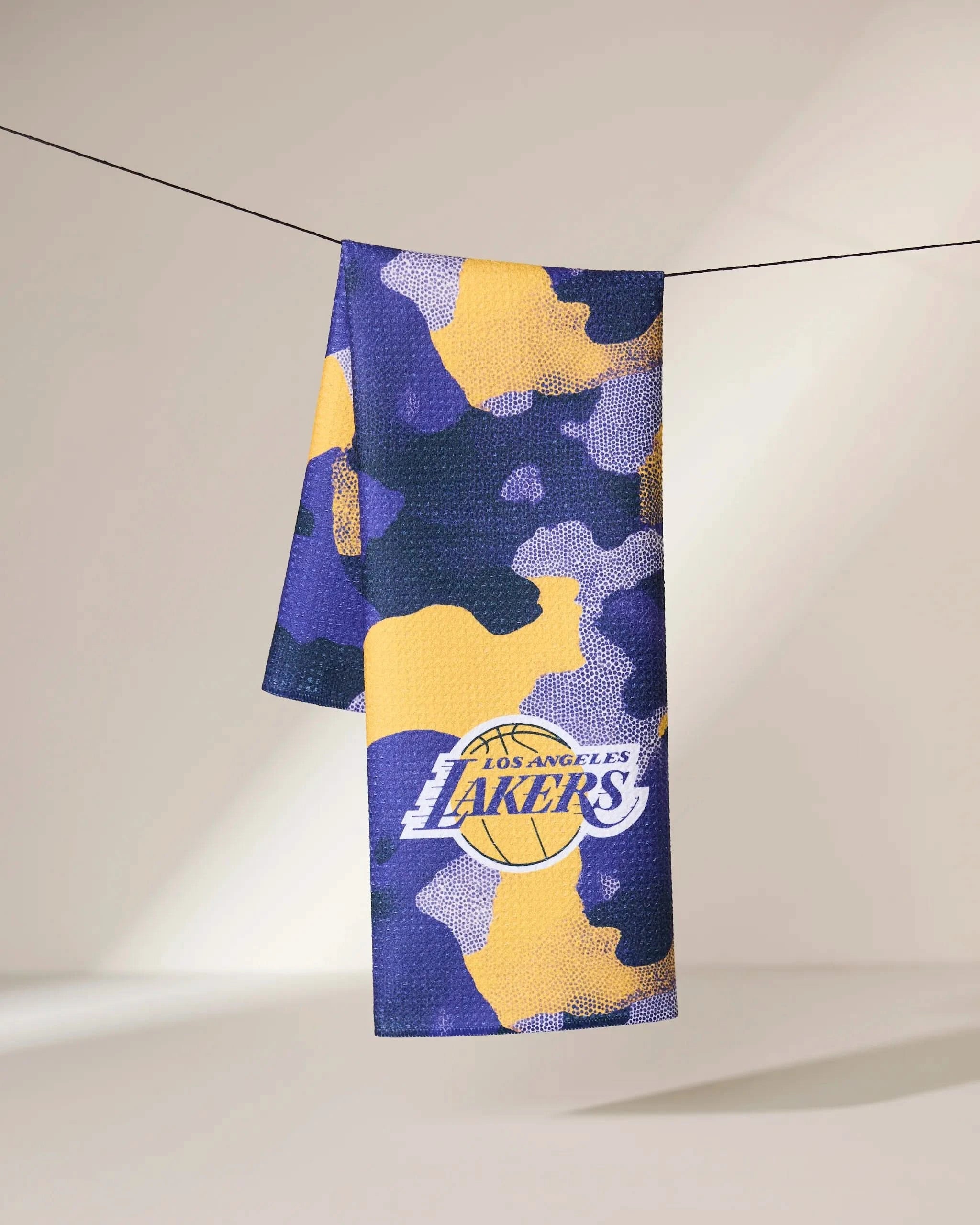 The Geometry Lakers Camo towel, showcasing a purple, yellow, and black camo design with the Los Angeles Lakers logo, is draped over a suspended black string against a soft beige background.