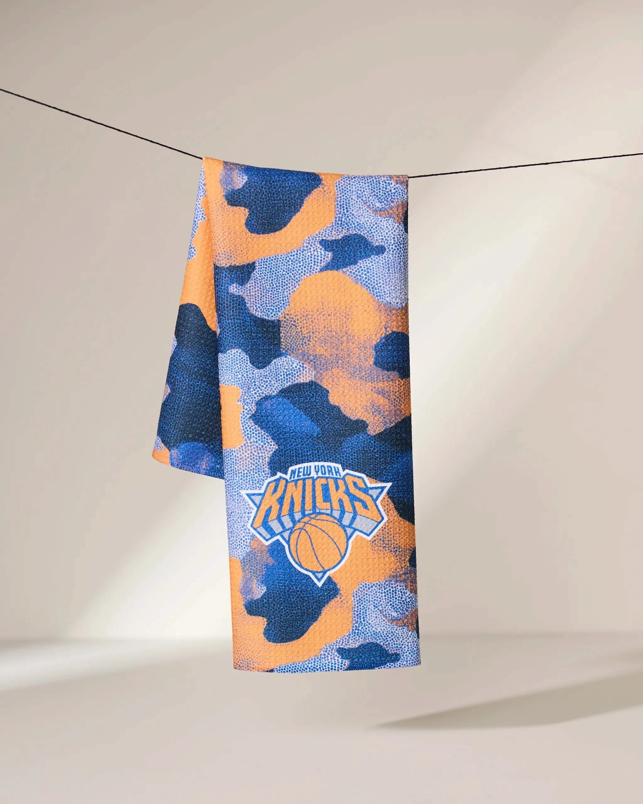 The Geometry Knicks Camo towel, featuring a blue, orange, and white camouflage pattern with the New York Knicks logo in the center, hangs on a line against a neutral, softly lit background.