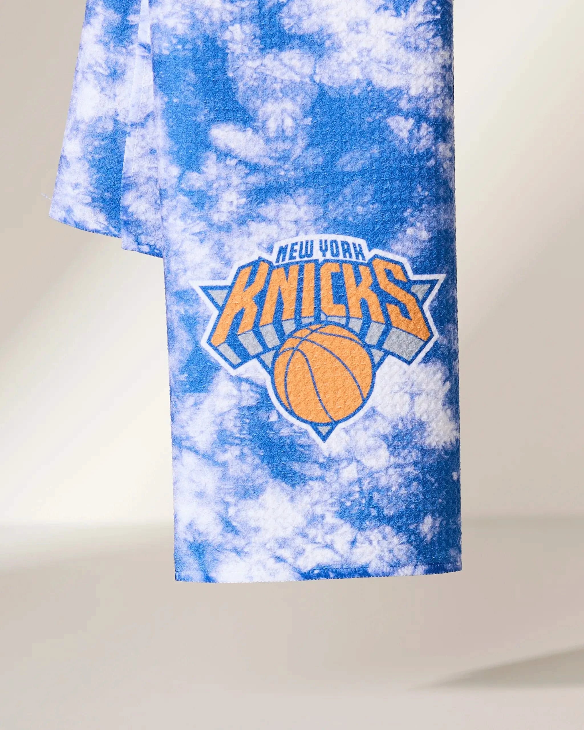 The Geometry Knicks Flash towel is a blue and white tie-dye design featuring the New York Knicks logo with an orange basketball and bold team name.