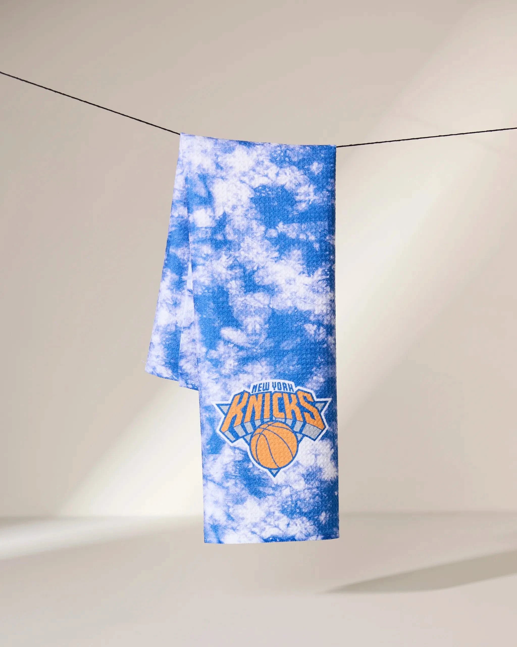 The Geometry Knicks Flash towel, featuring a blue and white tie-dye design with the New York Knicks logo, hangs over a black wire against a softly lit neutral background.