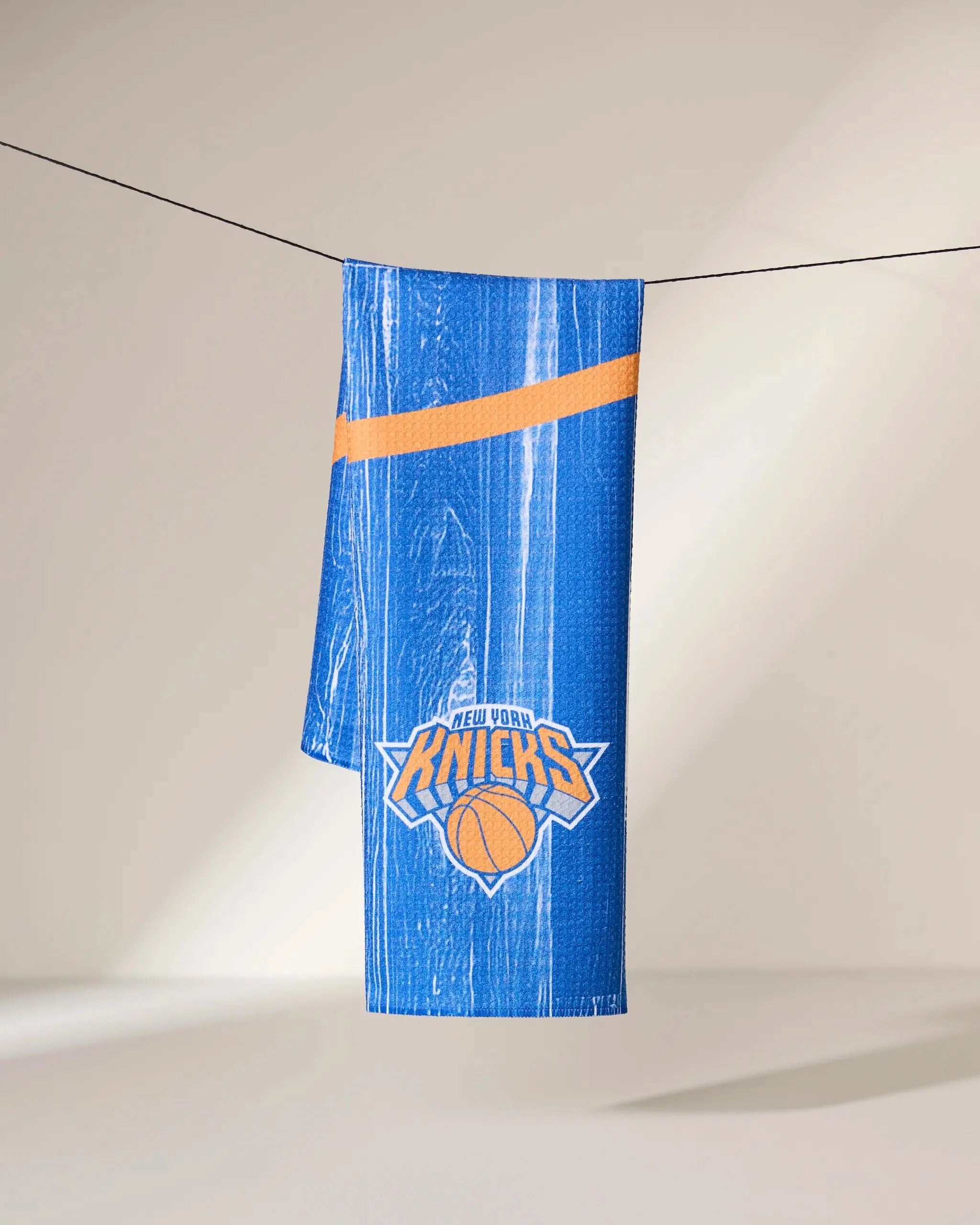 A Geometry Knicks Zone towel, featuring the New York Knicks logo and blue with an orange stripe, hangs draped over a clothesline against a neutral background.