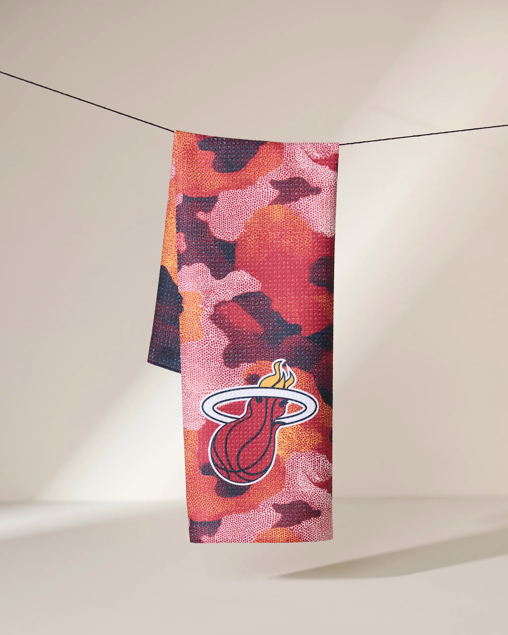 The Heat Camo towel by Geometry, featuring a red, pink, and black camouflage pattern with a Miami Heat logo, hangs on a string against a light background.