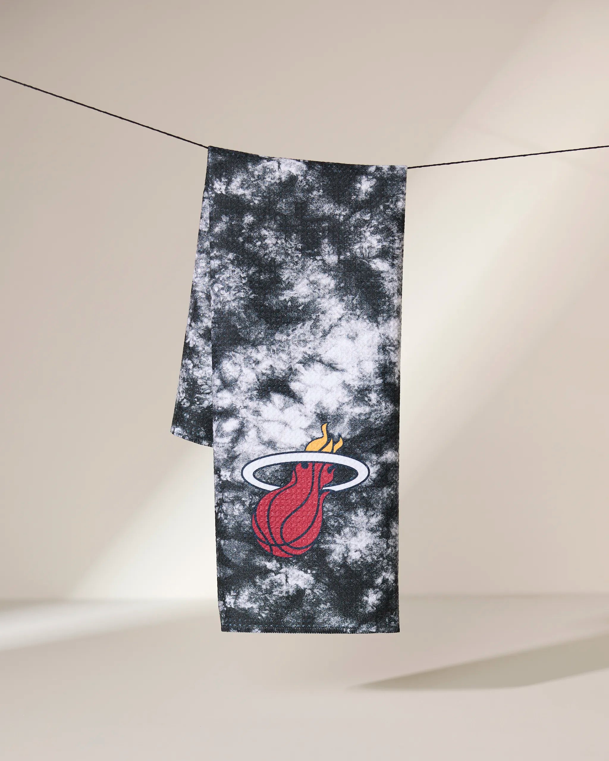 The Geometry Heat Flash towel, featuring a black and white tie-dye design with the Miami Heat logo, hangs over a thin line against a neutral background.