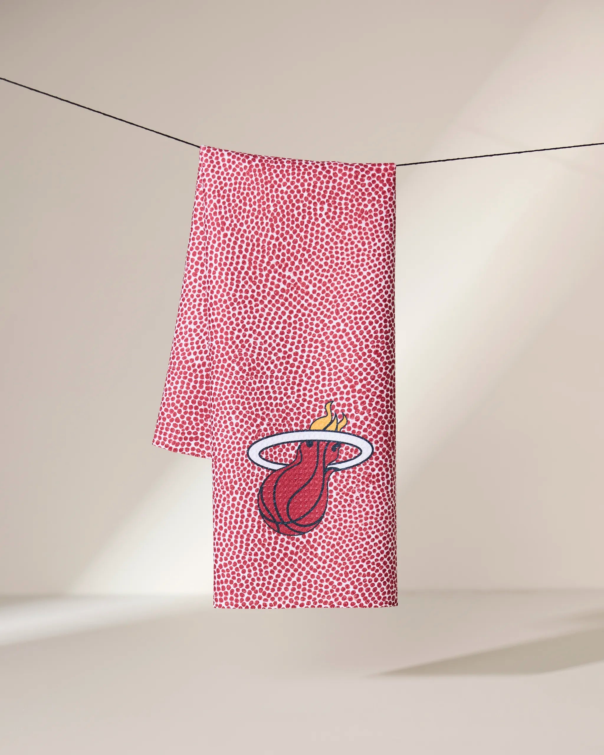 A red Geometry Heat Grip towel with a textured dot pattern hangs on a line. The Miami Heat logo appears near the bottom, set against a softly lit neutral background.