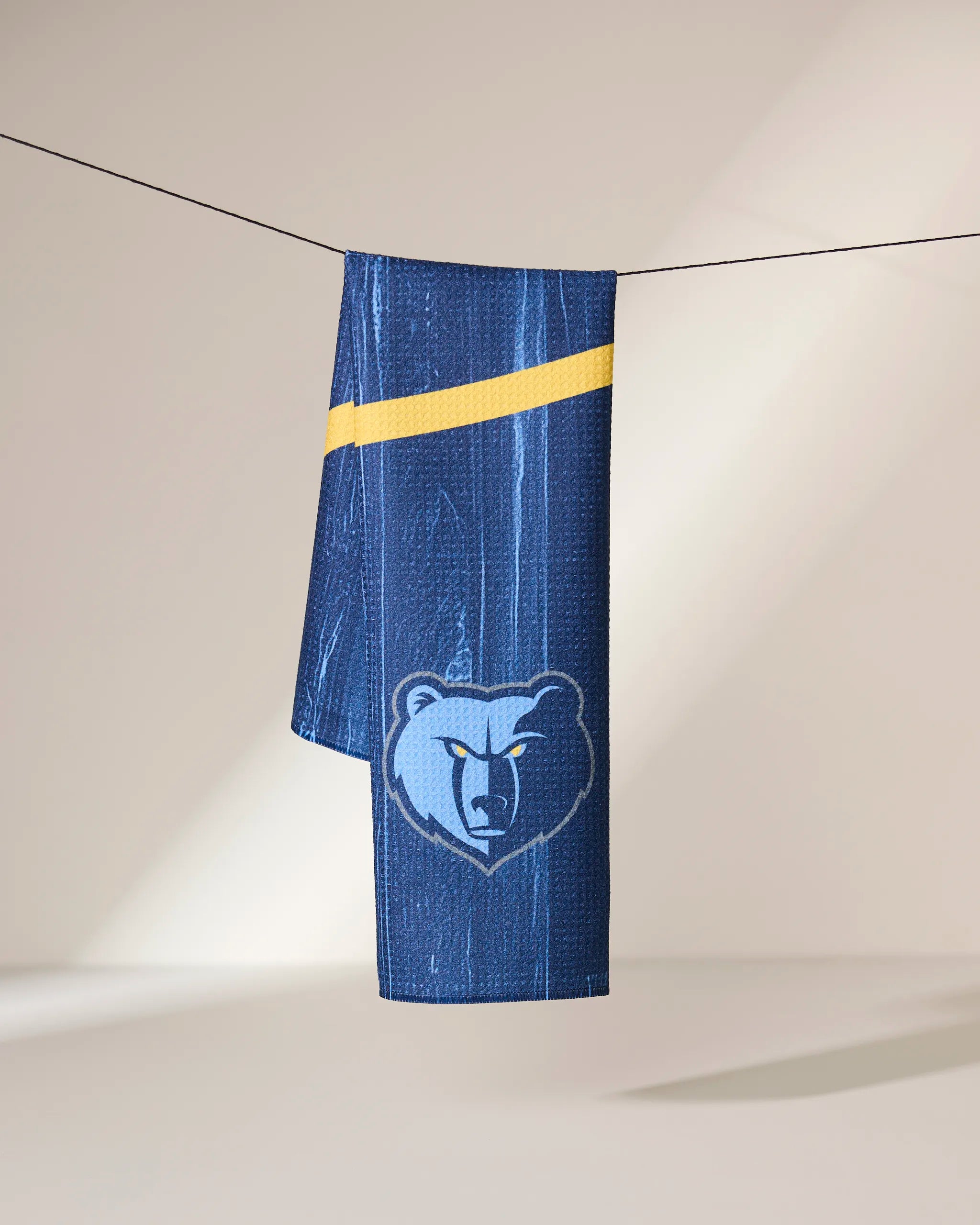 The Grizzlies Zone towel by Geometry, featuring a blue background, yellow diagonal stripe, and stylized bear head logo, hangs on a clothesline with a neutral backdrop.